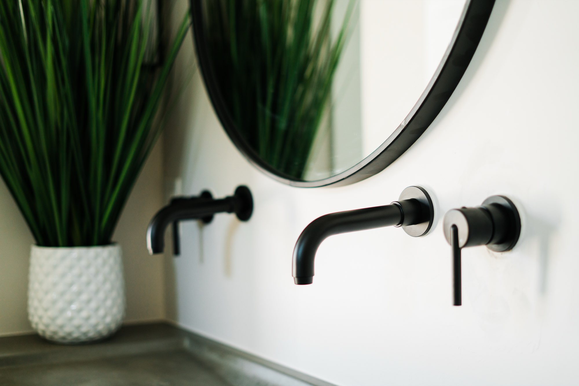 Close view of wall-mounted matte black faucets and the edge of a round black mirror