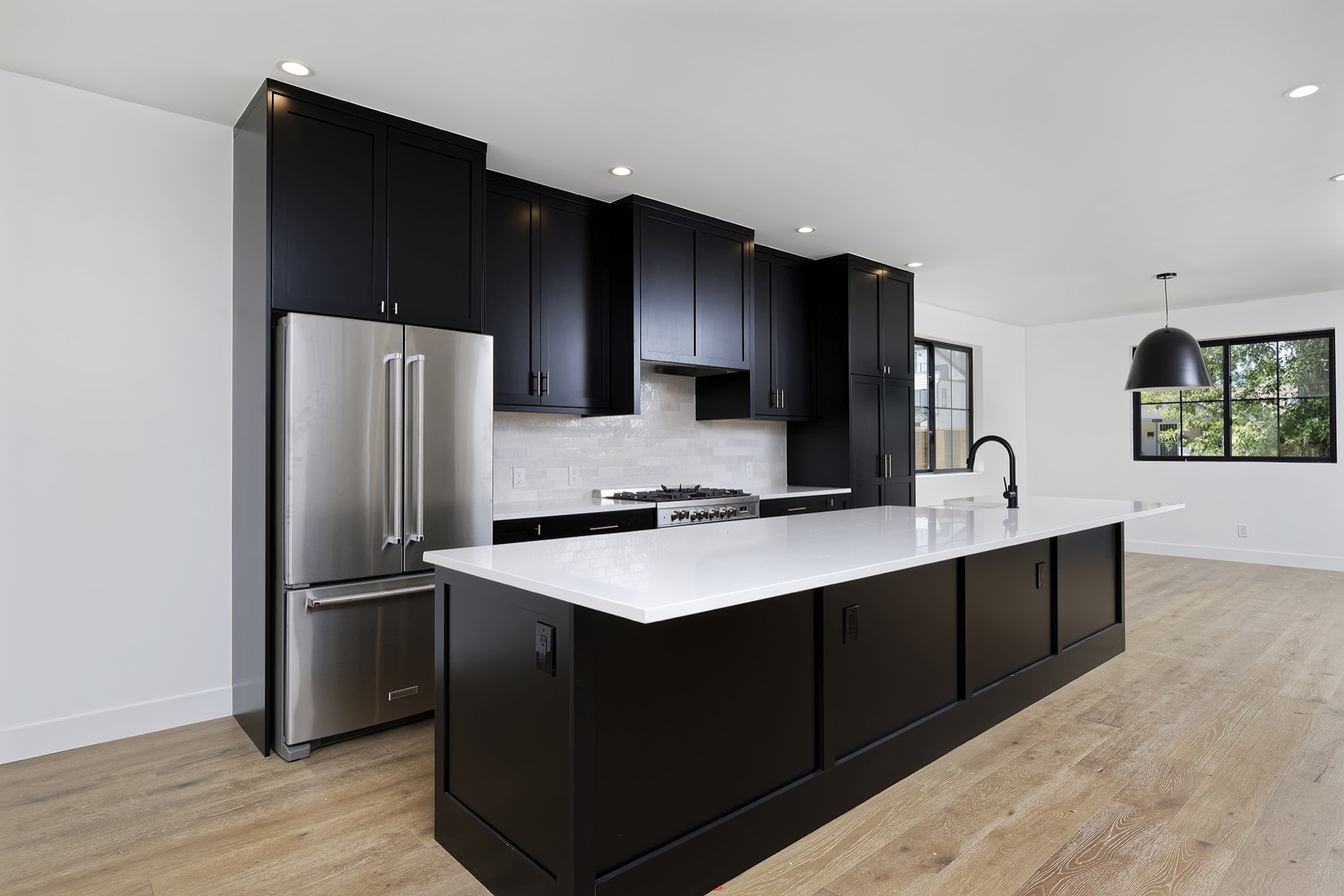 Wide view of the kitchen with black shaker cabinets, a long white island, stainless refrigerator and light wood floors