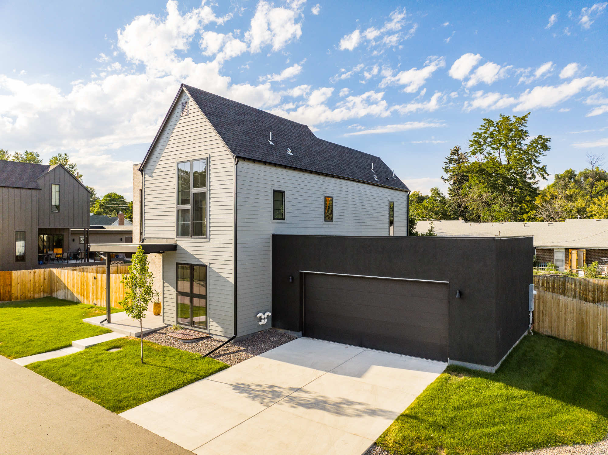 Corner view of the new Wheat Ridge custom home with a gray gable, dark shingle roof, dark two-car garage and a young tree out front
