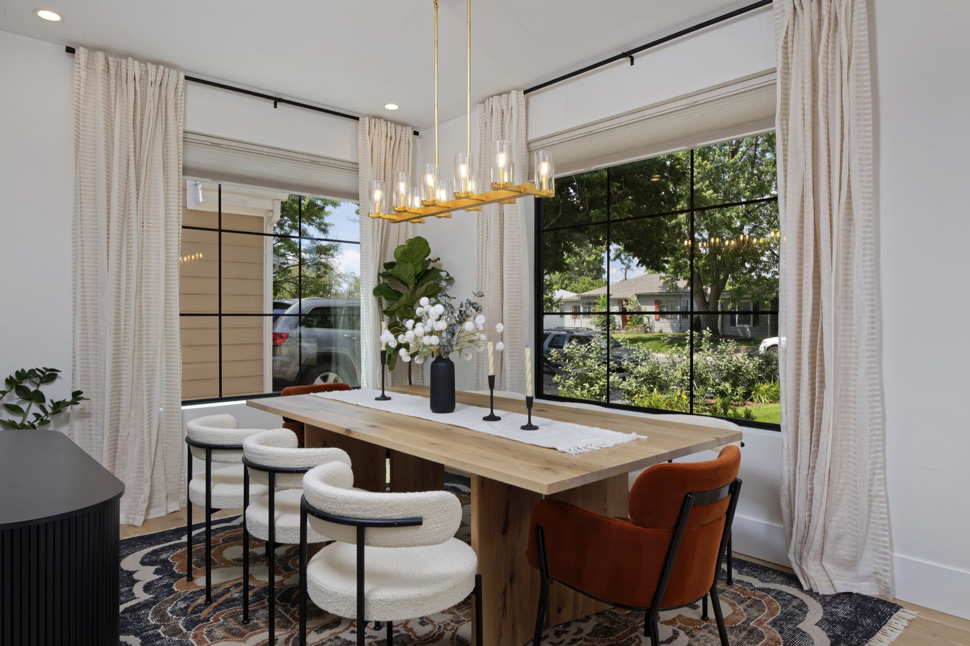 Dining room with a long wood table, brass chandelier, black-framed corner windows and linen drapes