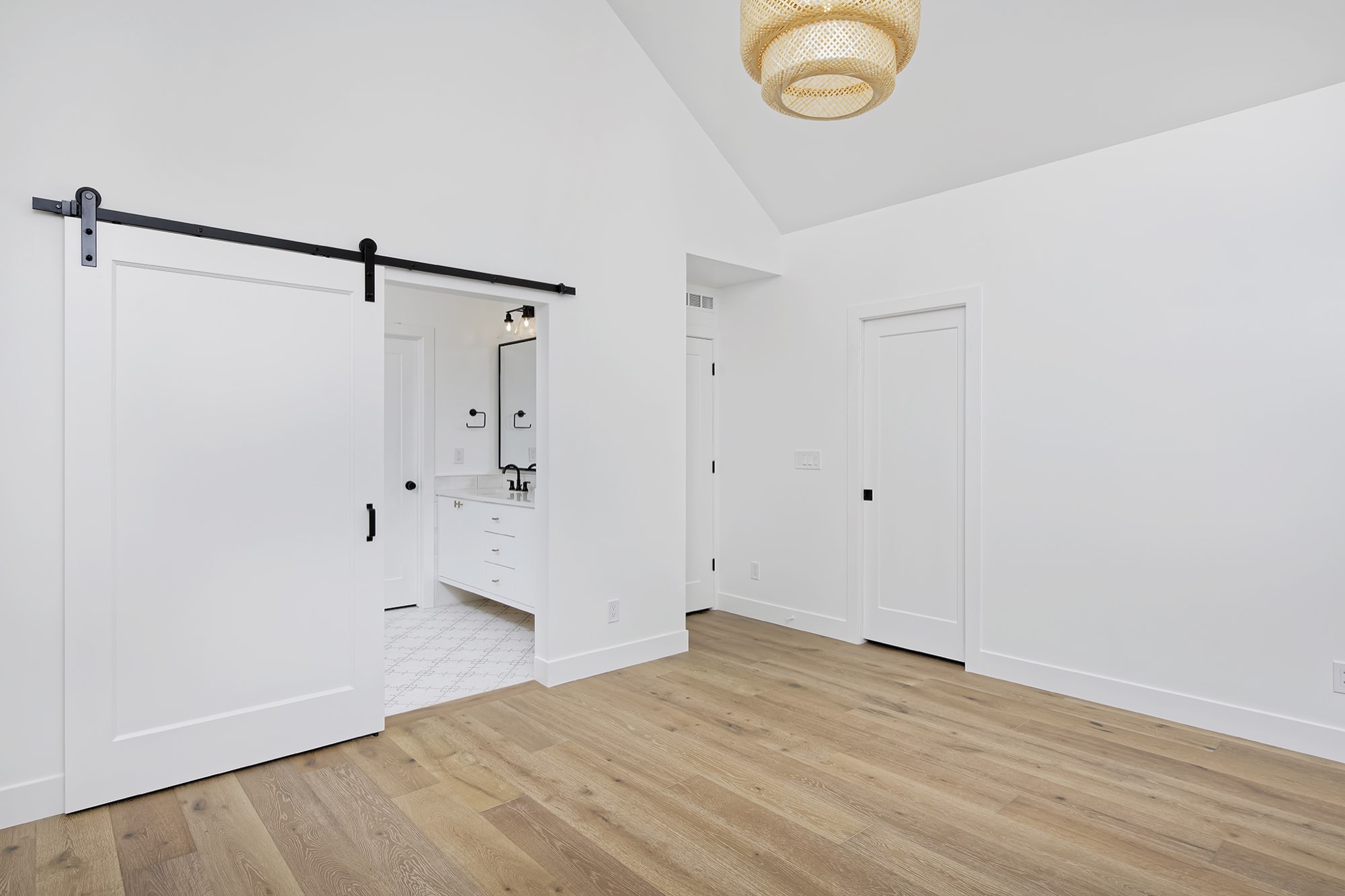 Bedroom with a vaulted ceiling, woven pendant, white barn door to the bath and light wood floors