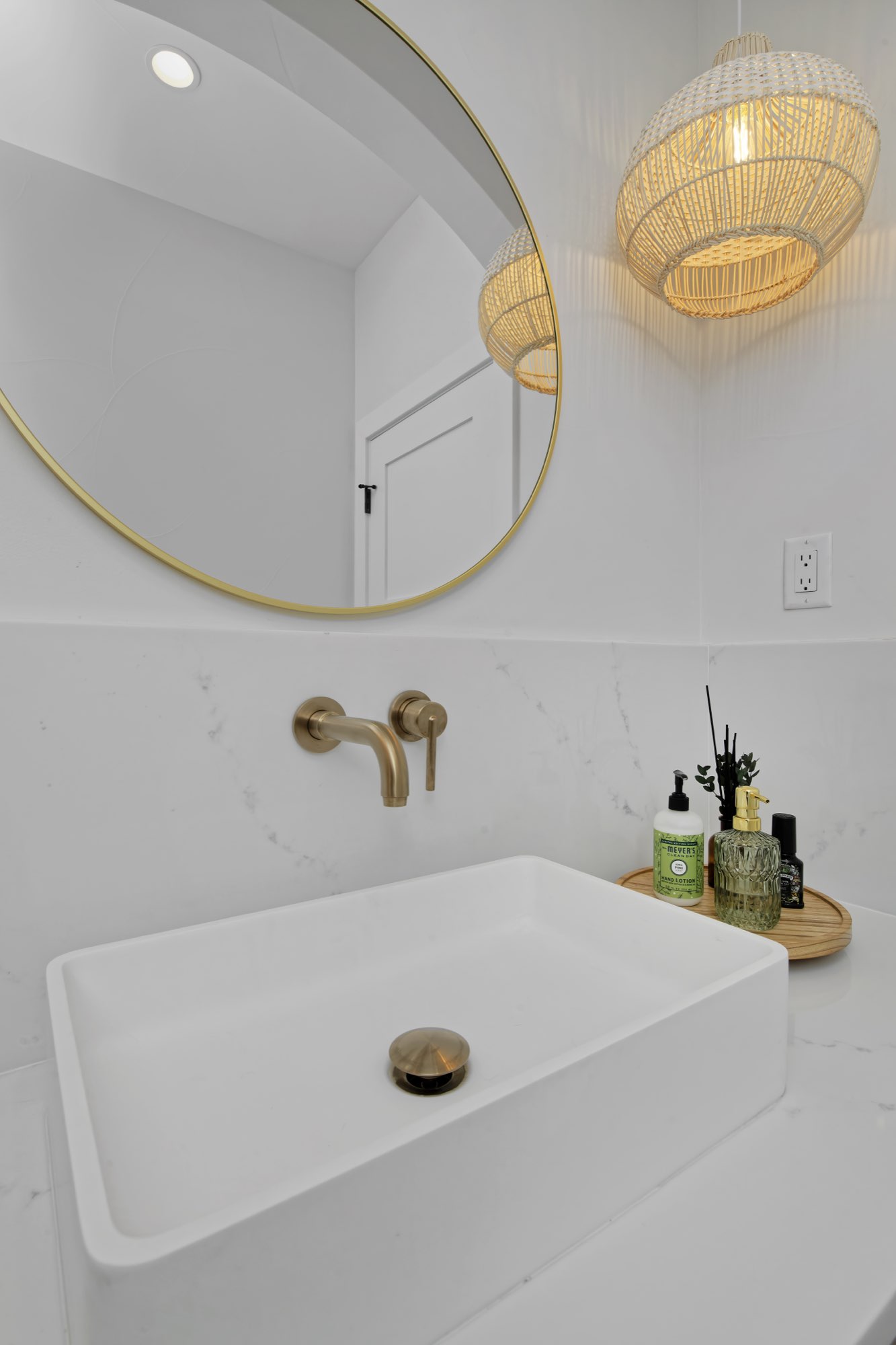 Vessel sink with a wall-mounted brass faucet, round brass mirror and a woven pendant light