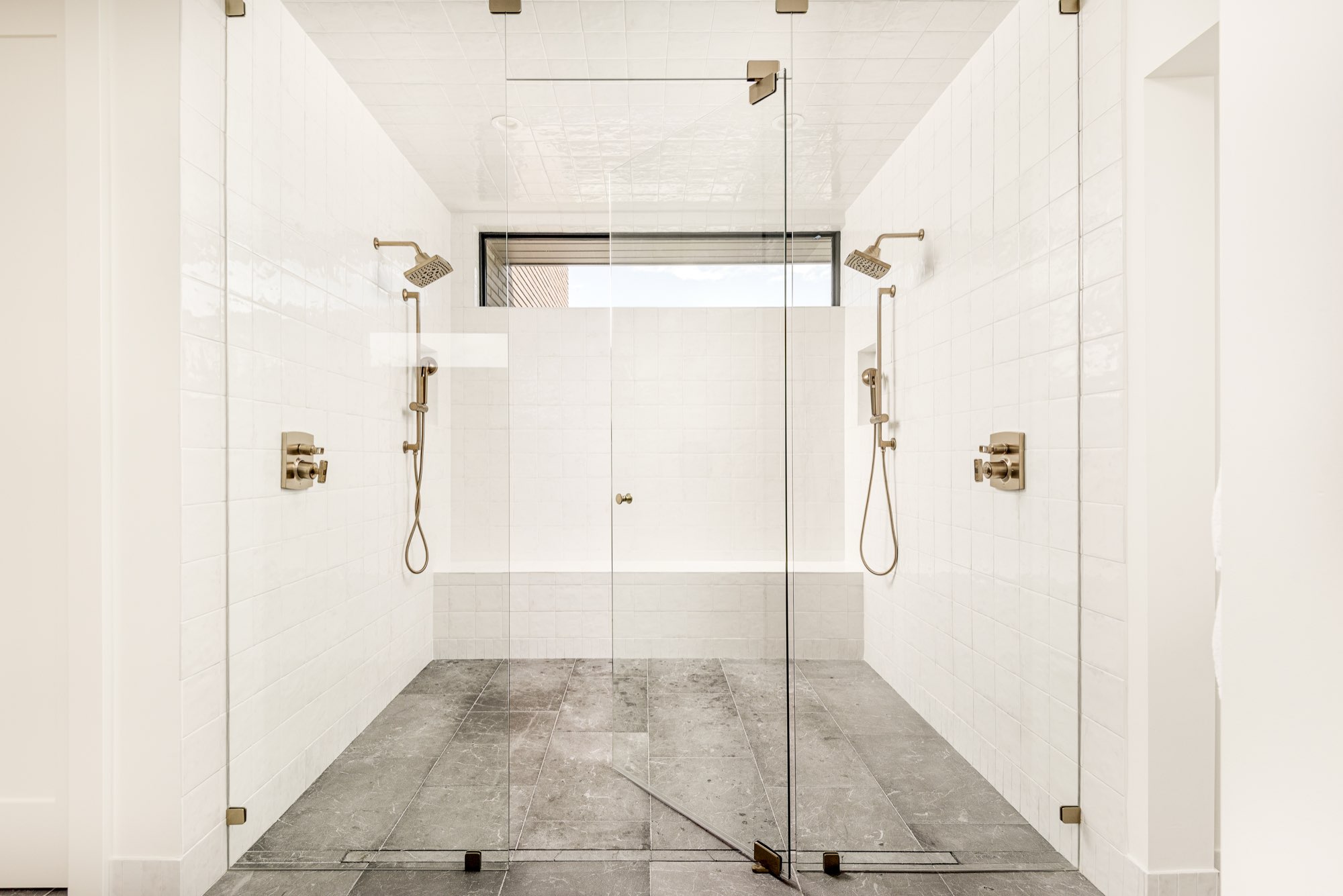Double walk-in shower behind clear glass with two shower heads, brass fixtures and white tile