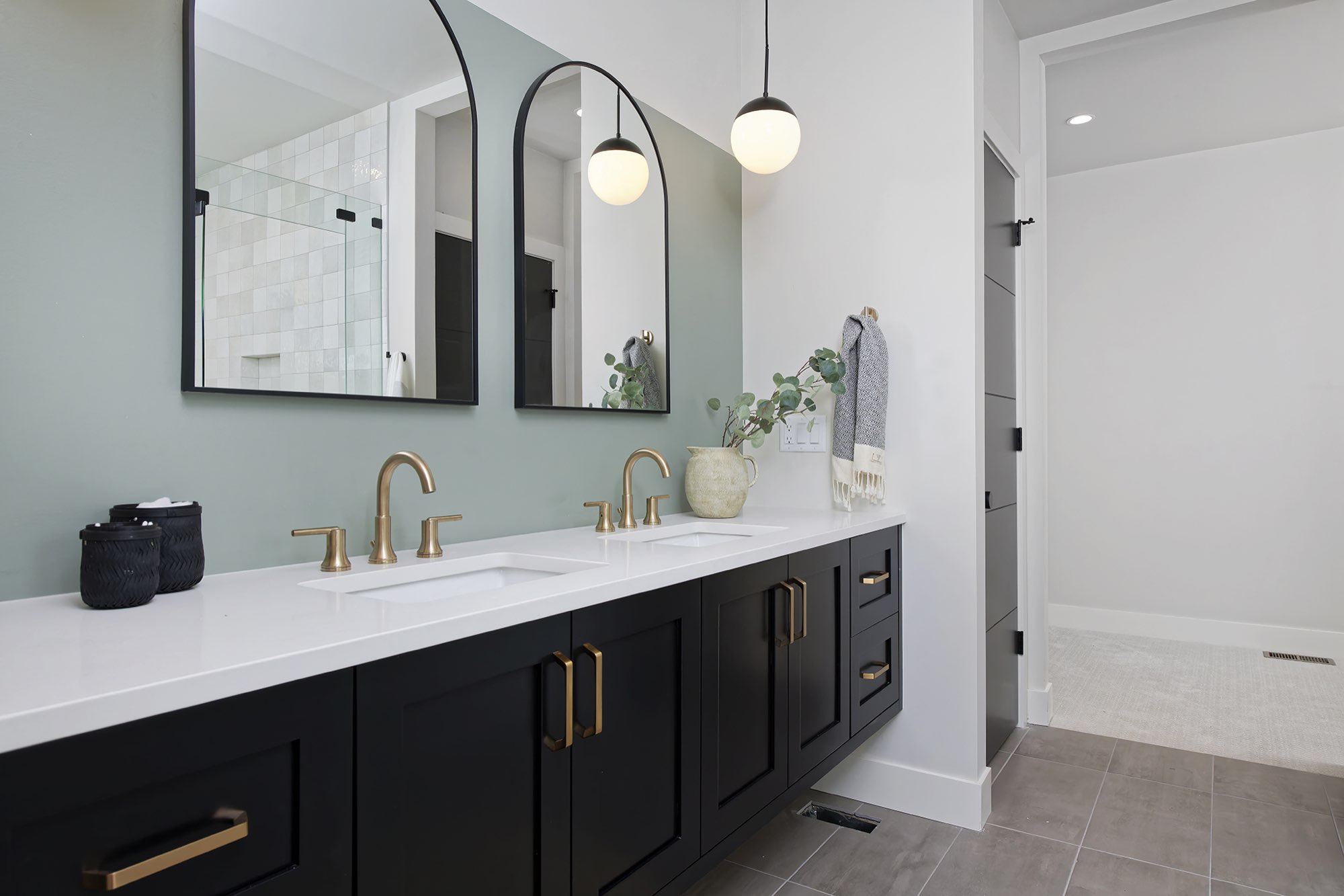 Long black double vanity with brass faucets and pulls under two arched black-framed mirrors against a sage wall