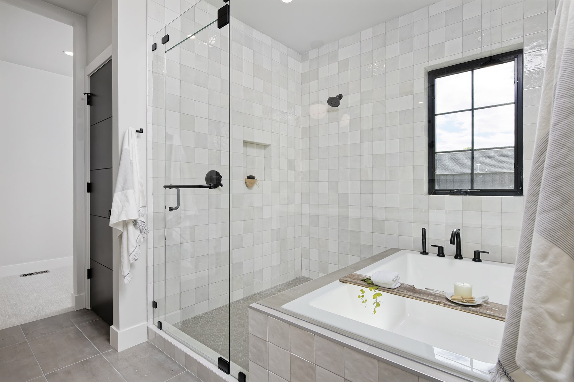 Primary bathroom with a glass shower and a drop-in soaking tub in soft gray square tile under a black-framed window