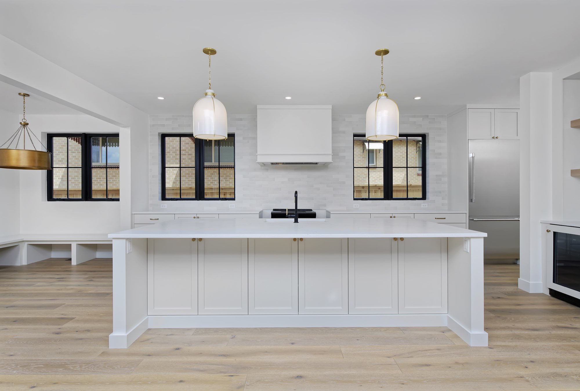 Front of the white kitchen island with two brass-trimmed pendants, black-framed windows and a hood over the range