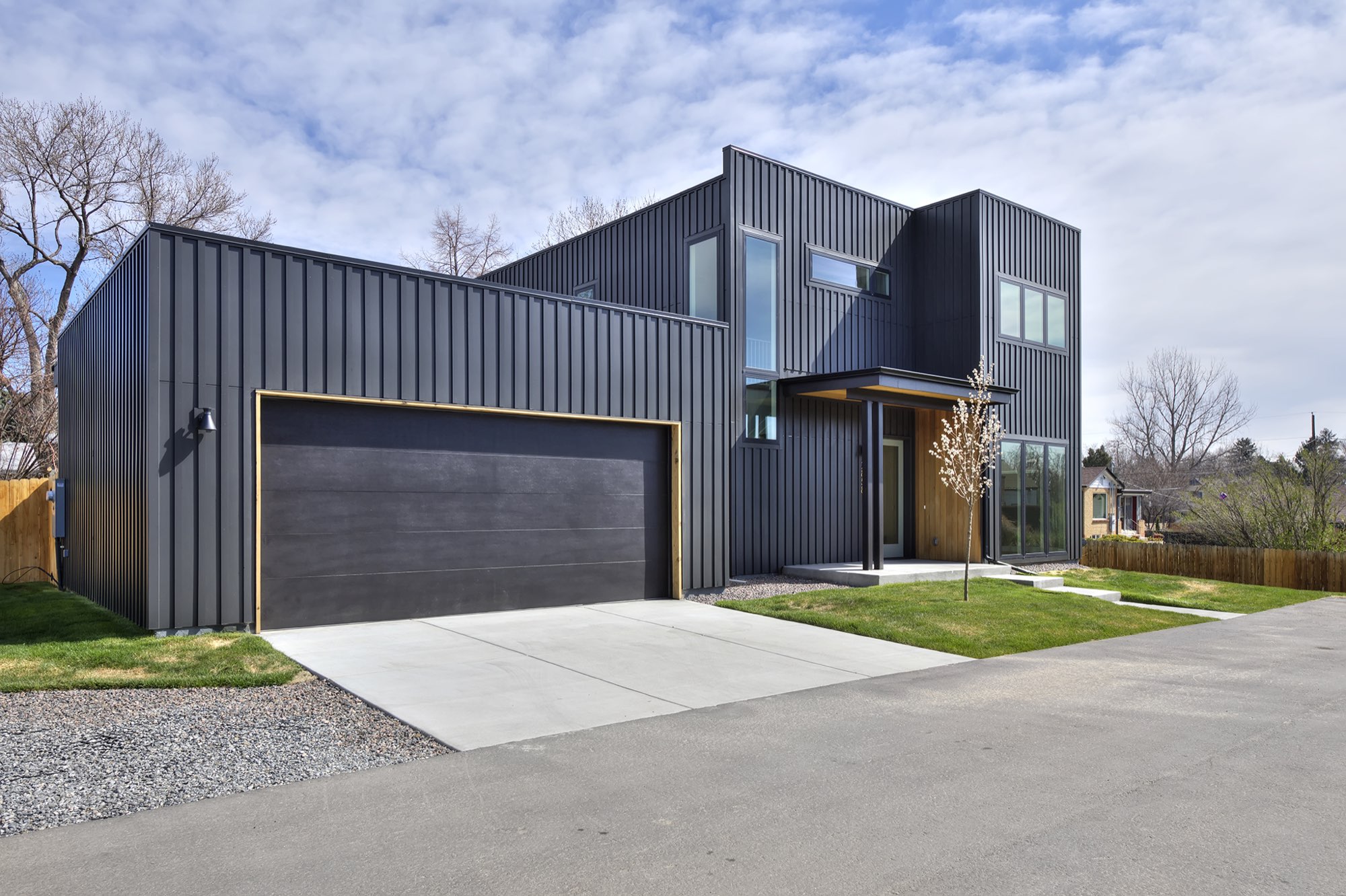 Two-car garage with a dark paneled door and wood trim beside the tall charcoal block, with a concrete drive and gravel edge