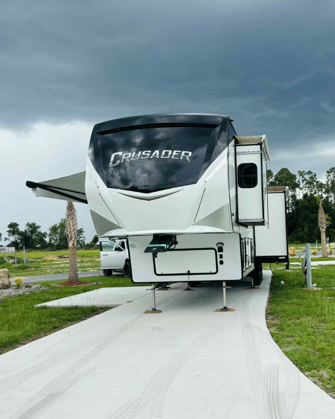 A fifth wheel RV backed into a spacious paved site with its awning extended under a dramatic Florida sky at Heritage Oaks RV Resort