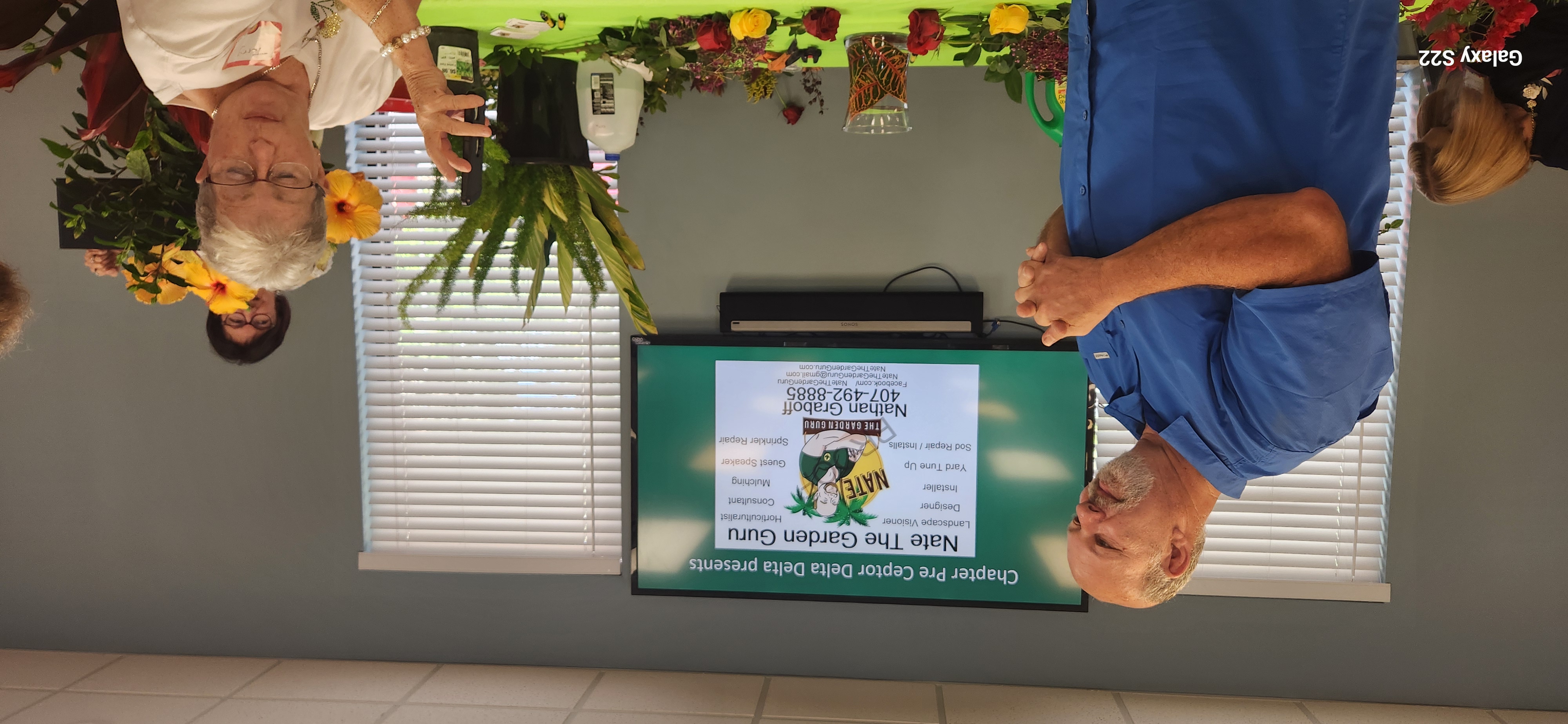 Nate the Garden Guru standing beside a presentation screen listing his landscaping services, with fresh-cut roses and tropical greenery on the table in front of him
