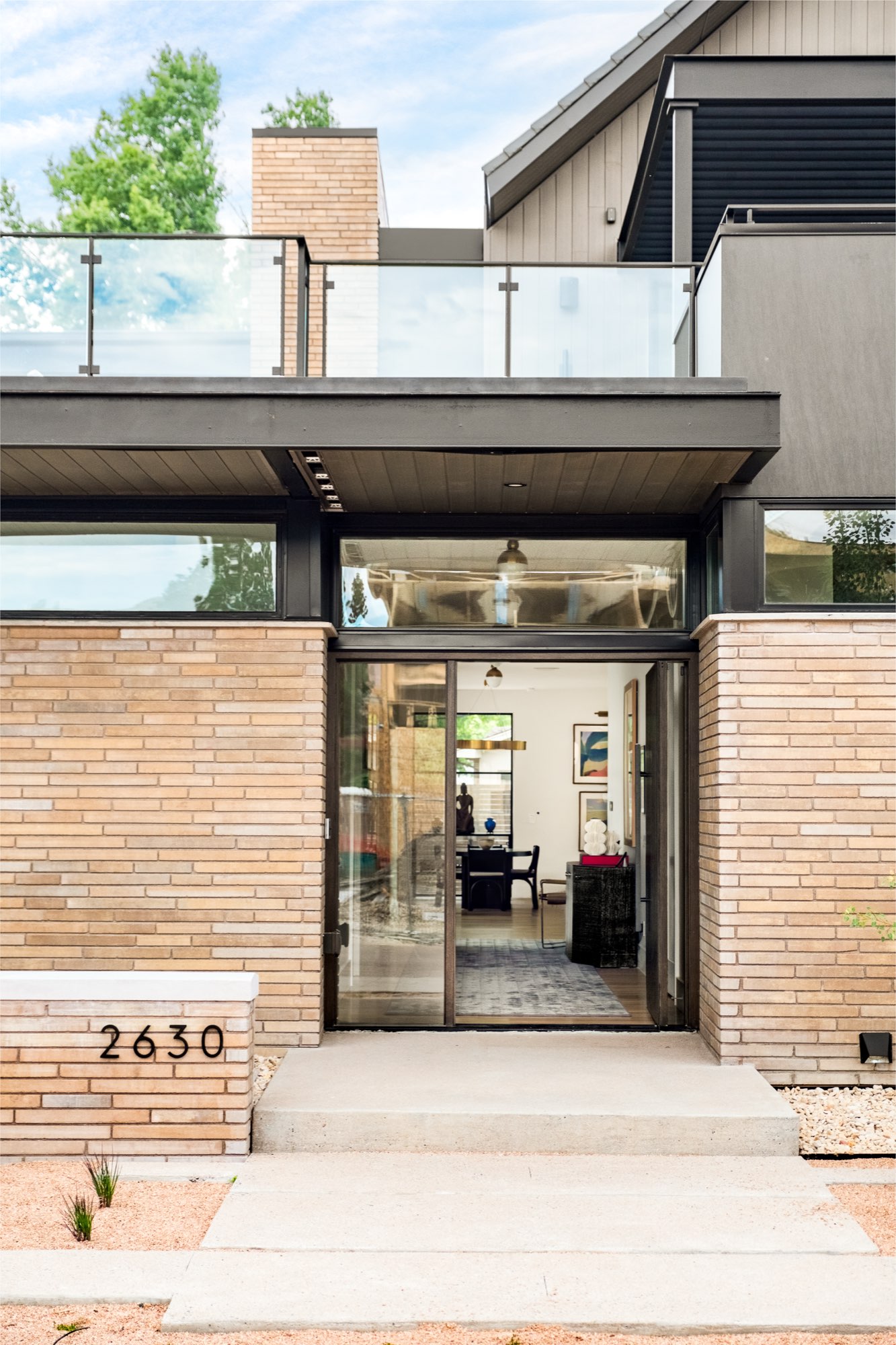 Glass front entry framed in tan brick, with the house number 2630 on a brick pier and a glass-railed terrace above