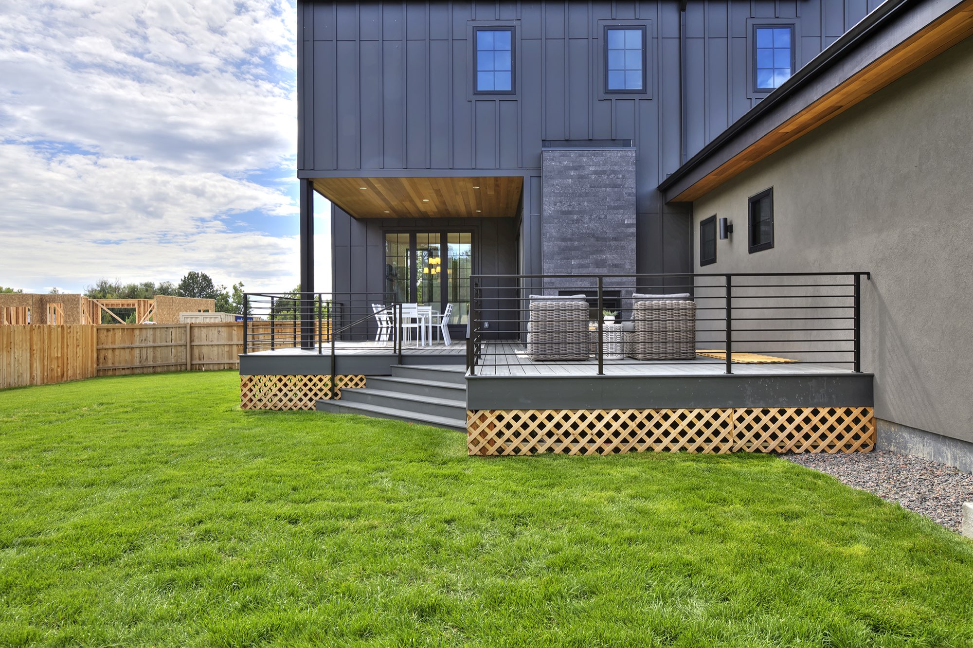 Back of the house from the lawn, with a covered deck, black railing, lattice skirt and stone fireplace chimney