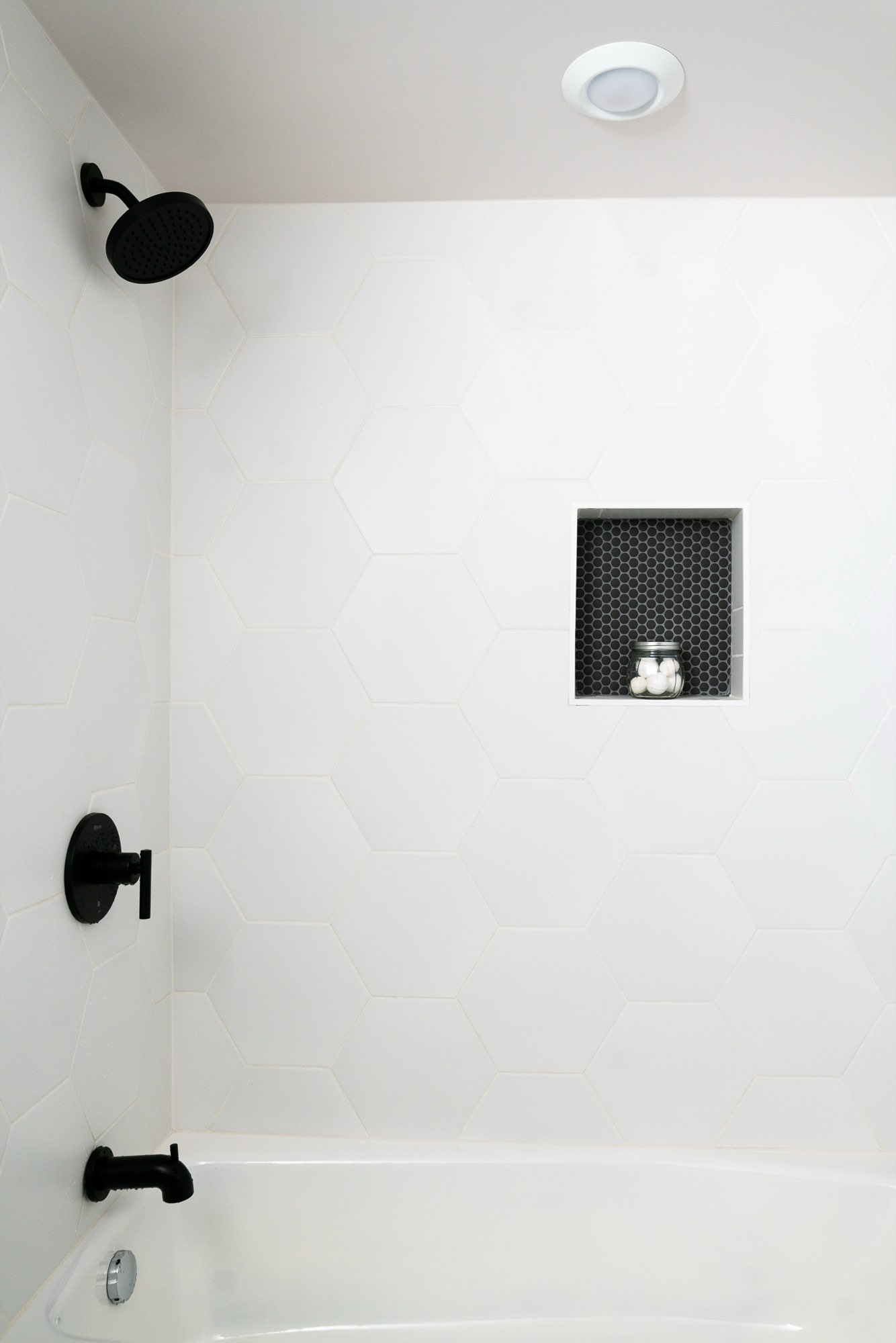 Shower and tub with large white hexagon tile, a black shower head and a small black tile niche