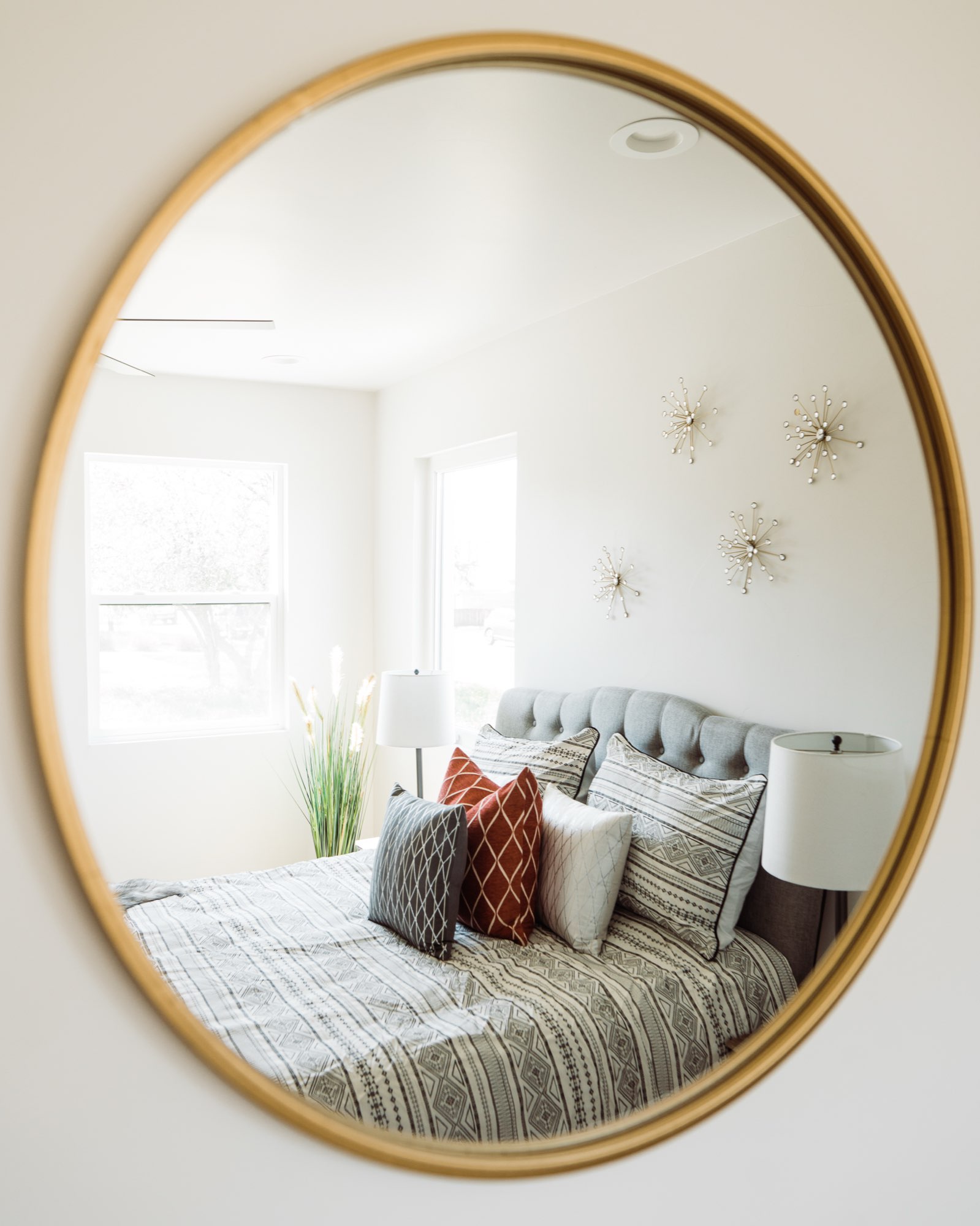 Round gold-framed mirror reflecting a bedroom with a tufted headboard and metal starburst wall decor