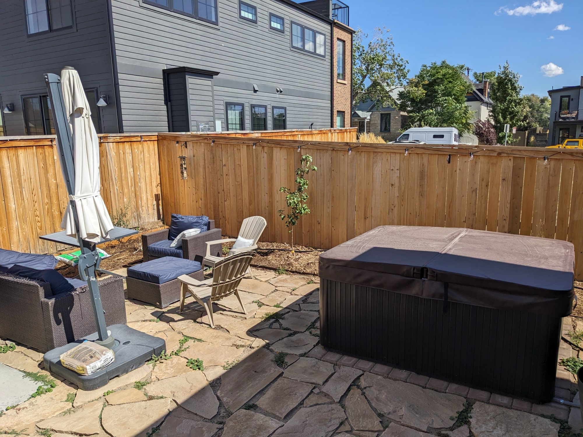 Backyard with a flagstone patio, outdoor seating, a covered hot tub and a wood fence on a sunny day