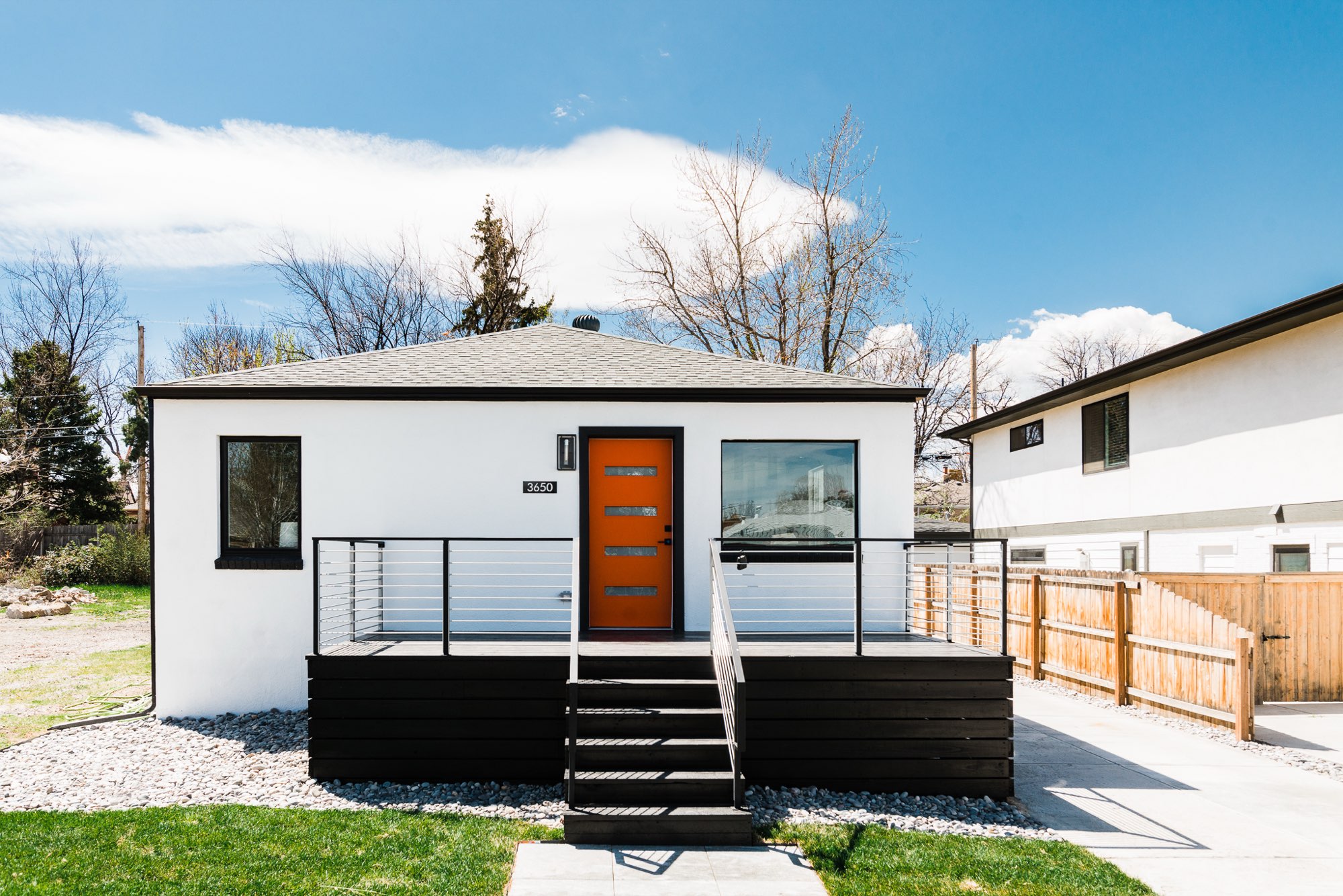Straight-on front of the white bungalow with an orange front door, black trim, a dark deck and a cable railing