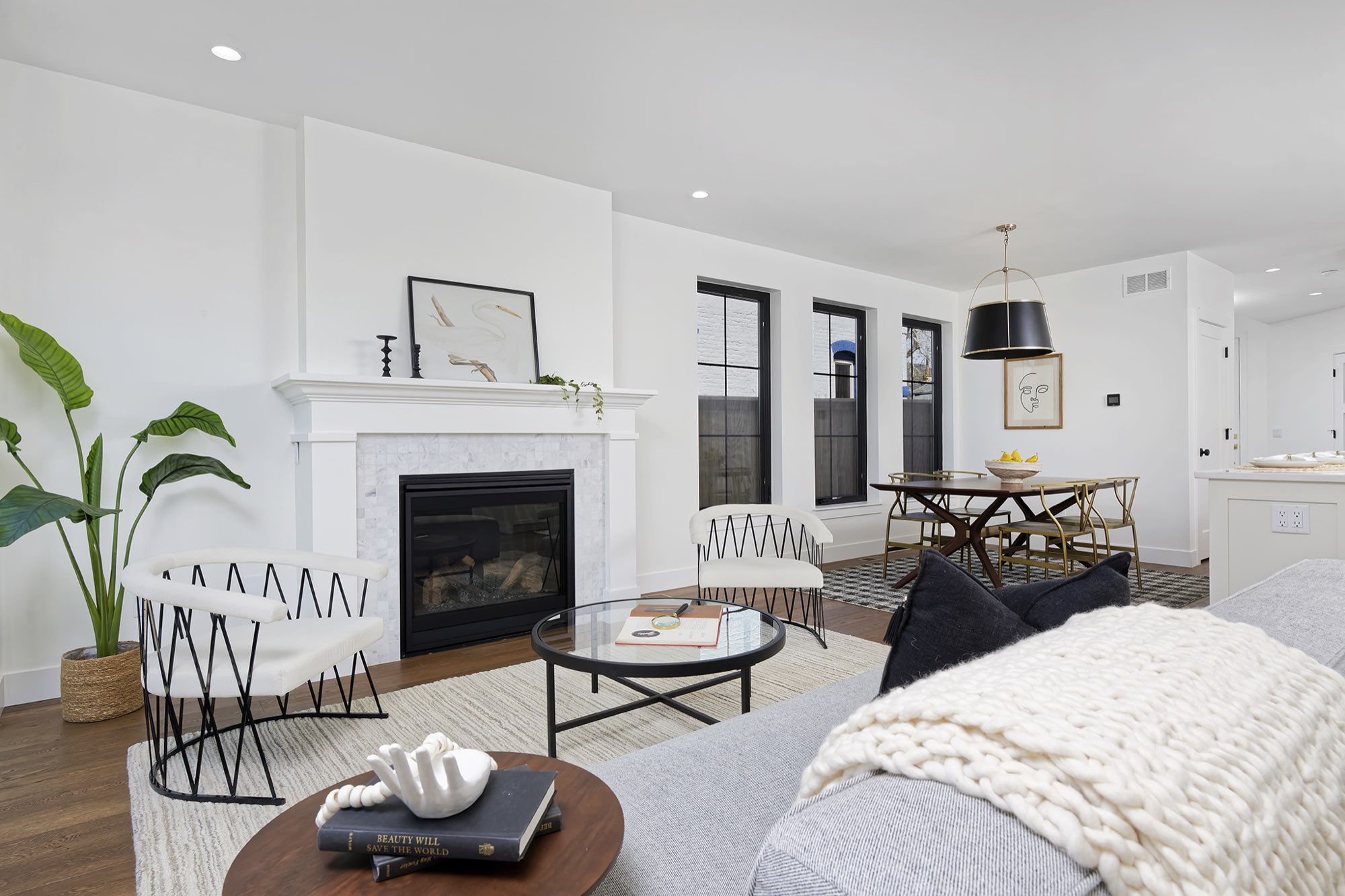 Open living and dining room with a white fireplace mantel, black-framed windows, wood floors and a black pendant over the dining table
