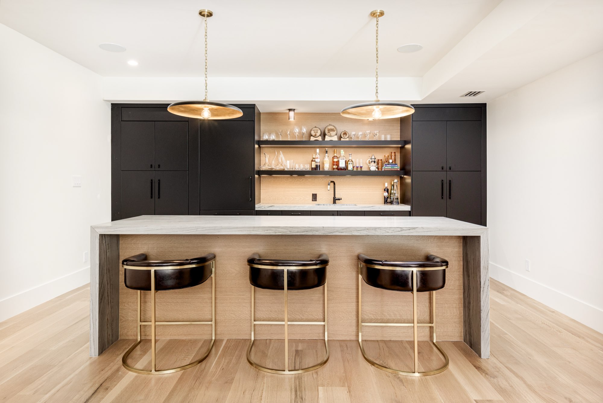 Bar with black cabinets, open shelves, a stone counter, three brass-framed stools and two brass pendants