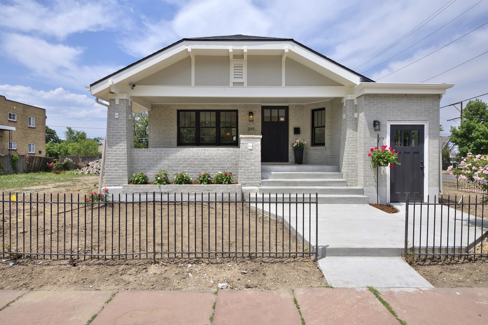 Straight-on front of the renovated bungalow with pale gray brick, a covered porch, black-framed windows and a black iron fence