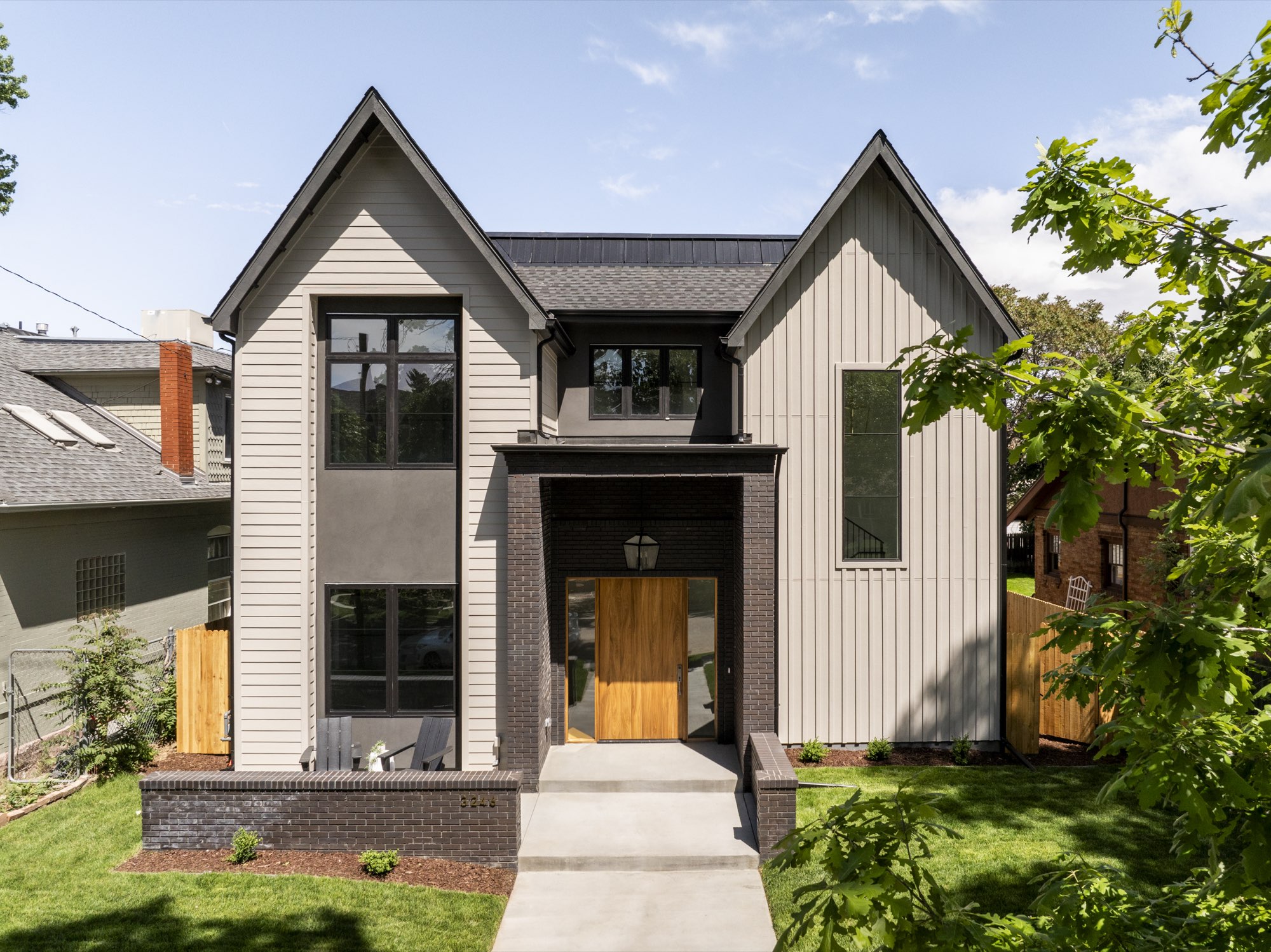 Front of the new custom home in Denver with two light siding gables, black-framed windows, a dark brick base and a wood front door