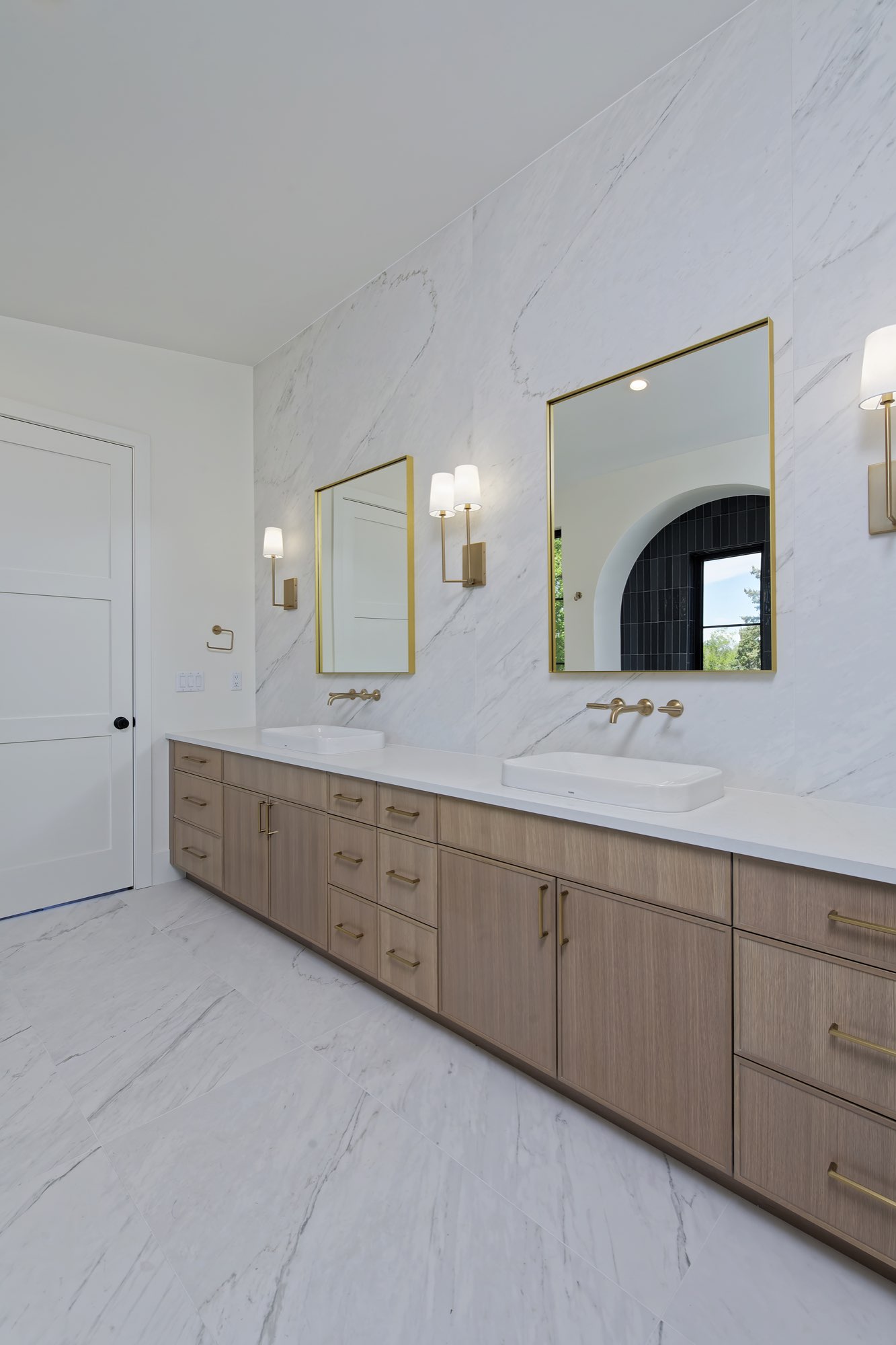 Long wood-tone double vanity with gold-framed mirrors, brass sconces and wall-mounted faucets on white veined stone