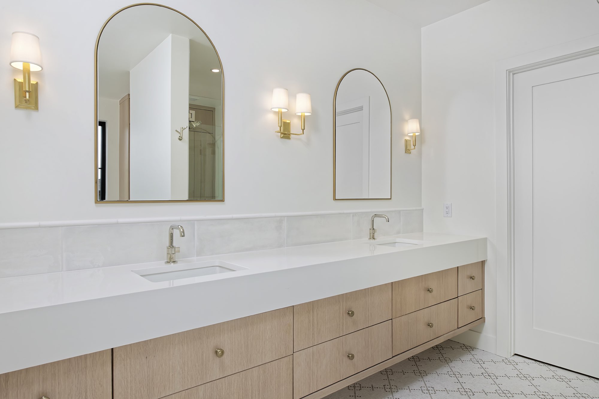 Primary bath double floating wood vanity with arched brass-framed mirrors, brass sconces and a patterned mosaic floor