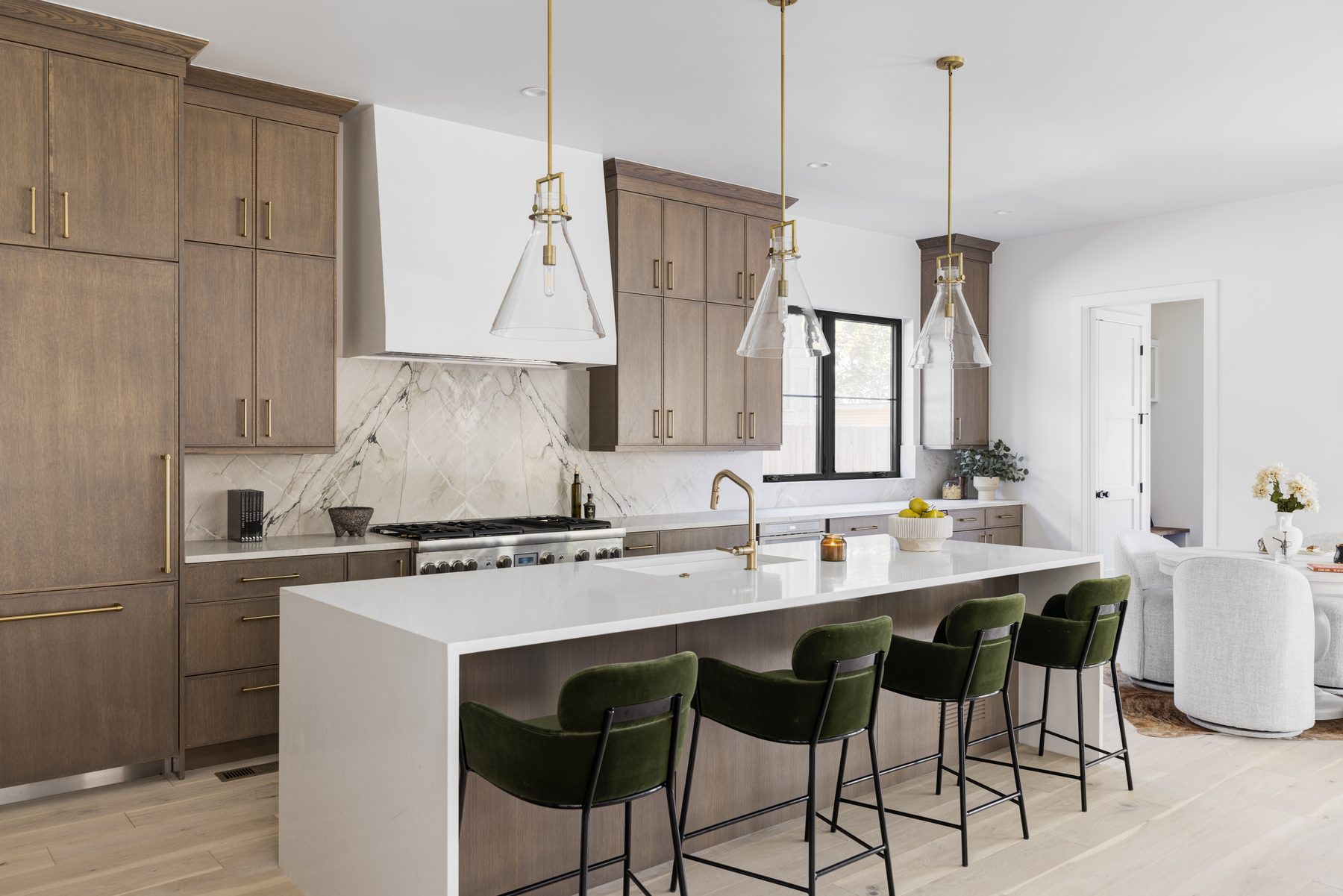 Kitchen with oak cabinetry, a white waterfall island with green velvet stools, marble backsplash and brass pendant lights