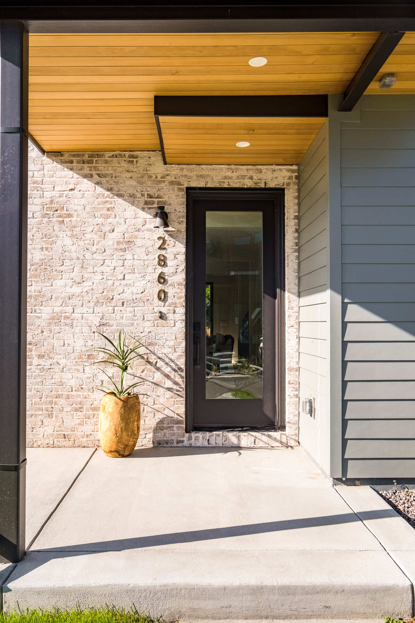 Front entry at 2860 Teller Street with pale brick, a black-framed glass door, a wood-plank soffit and house numbers