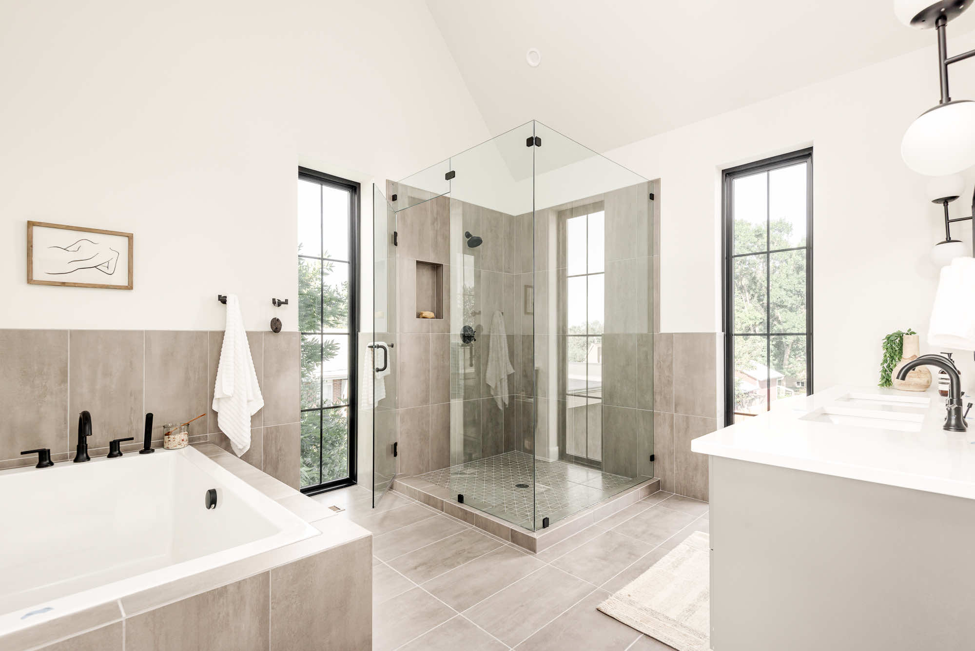 Primary bathroom with a corner glass shower, soaking tub and tall black-framed windows under a vaulted ceiling