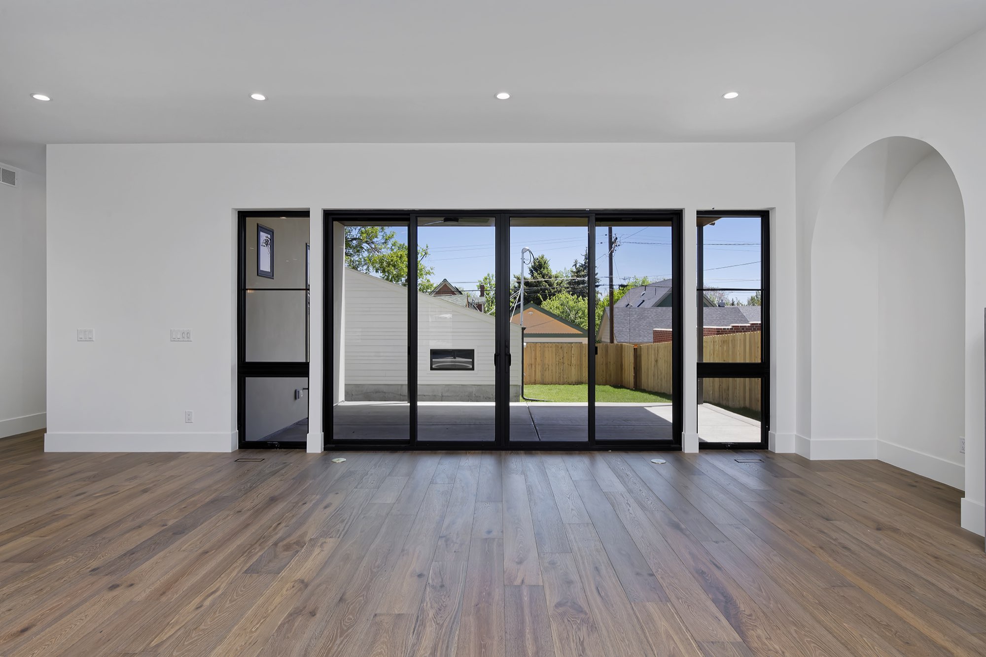 Living room with wide-plank wood floors, a wall of black-framed sliders and an arched niche, looking toward the back patio