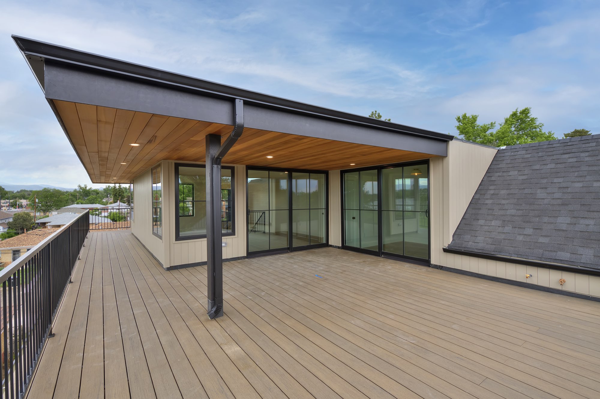 Wide rooftop deck under a wood-plank ceiling with recessed lights, black columns and railing, and black-framed sliders