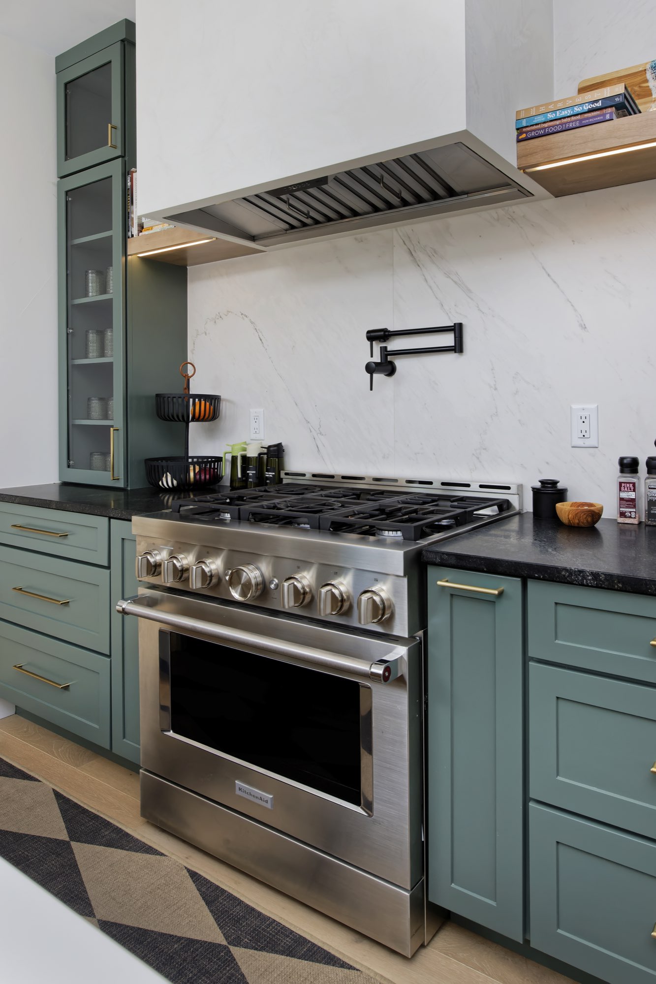 Stainless range with a white hood, black pot filler and sage green cabinets with brass pulls