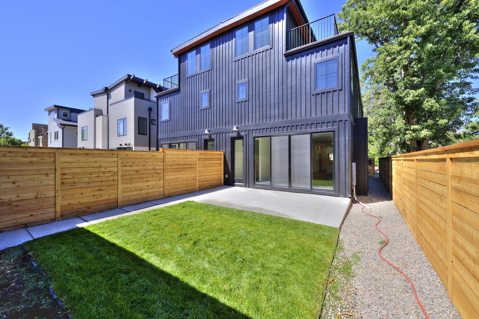 Rear of the duplex with black siding, sliding glass doors, concrete patio, lawn and wood privacy fence