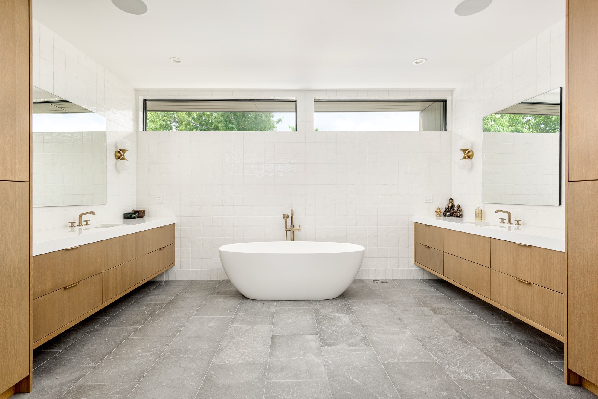 Primary bath with a freestanding tub between two floating wood vanities and a row of high windows