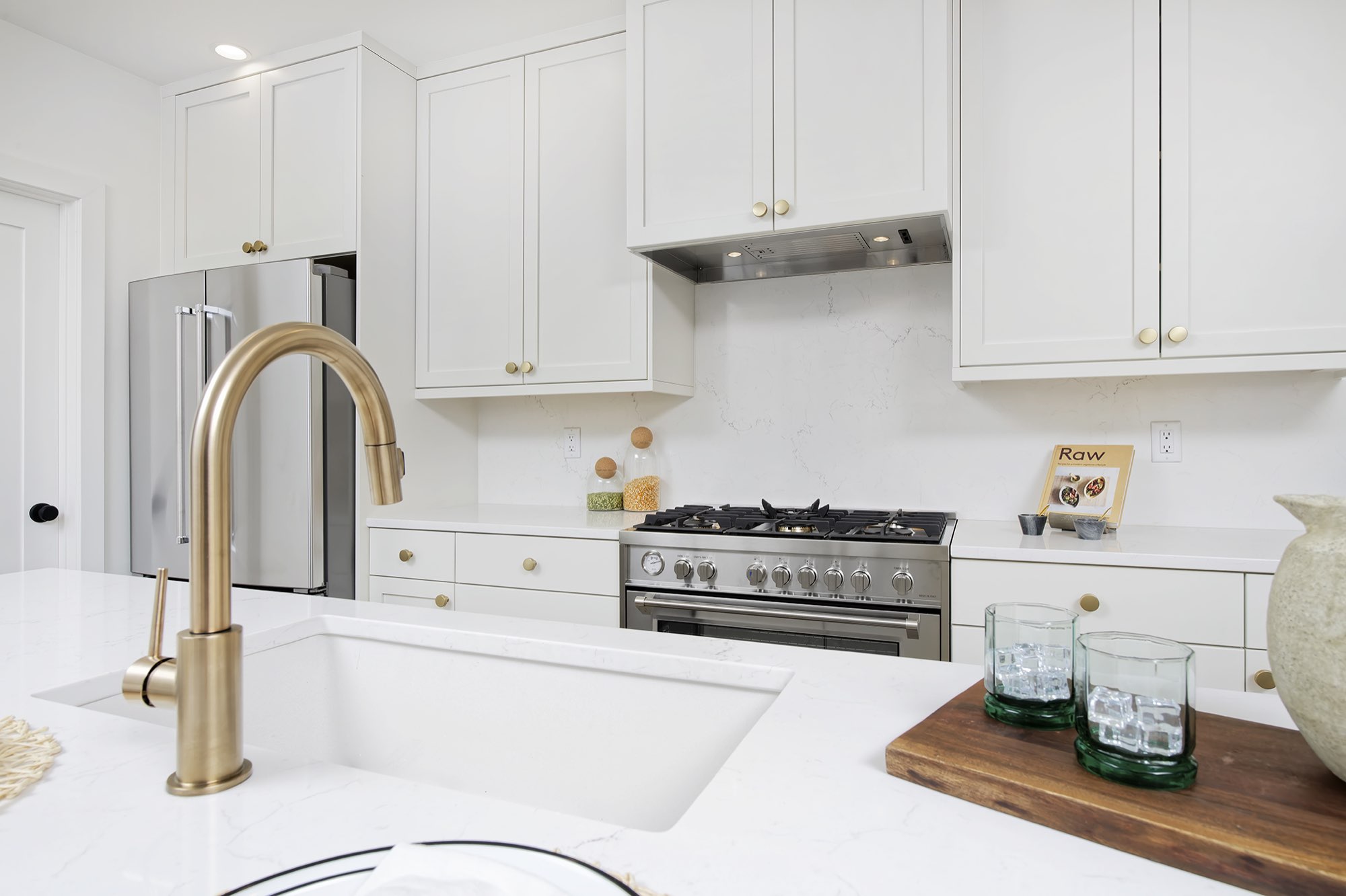 Kitchen sink with a brass faucet and white counters, with a gas range and white shaker cabinets behind