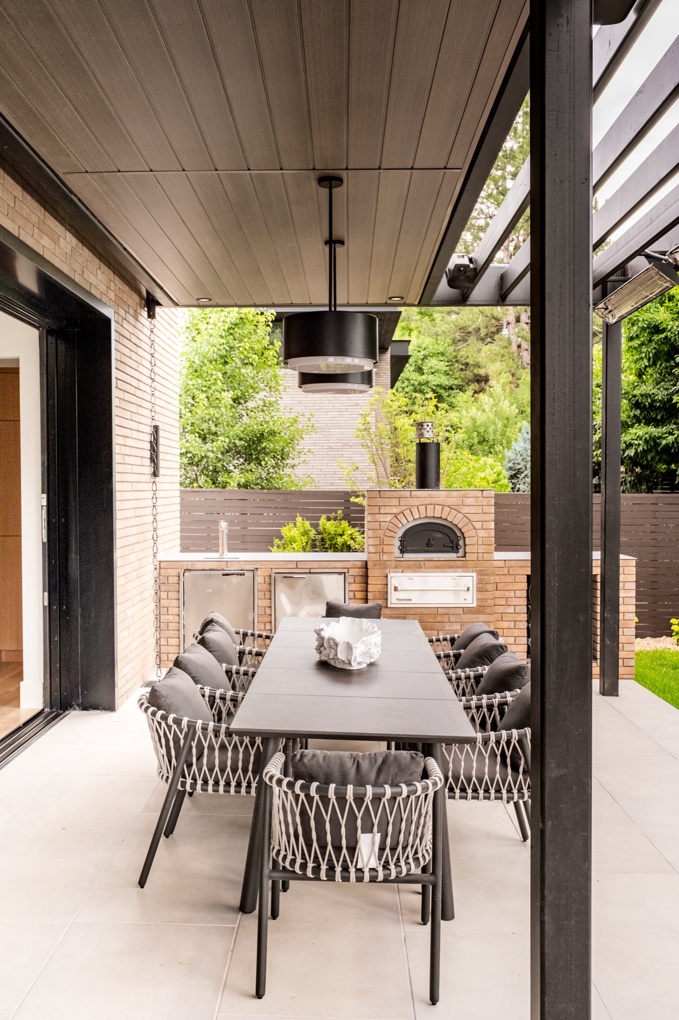 Covered patio with a long dining table, a brick oven and a black drum pendant