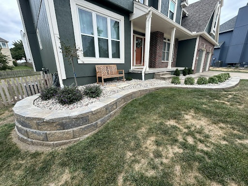Front yard landscape bed with a curved block retaining wall, white rock, shrubs, and a young tree in front of a home in Pleasant Hill, MO
