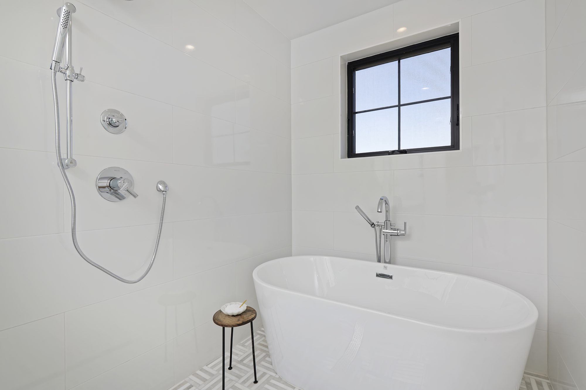 Primary bath with a freestanding white tub, tub filler, hand shower and a black-framed window over white tile