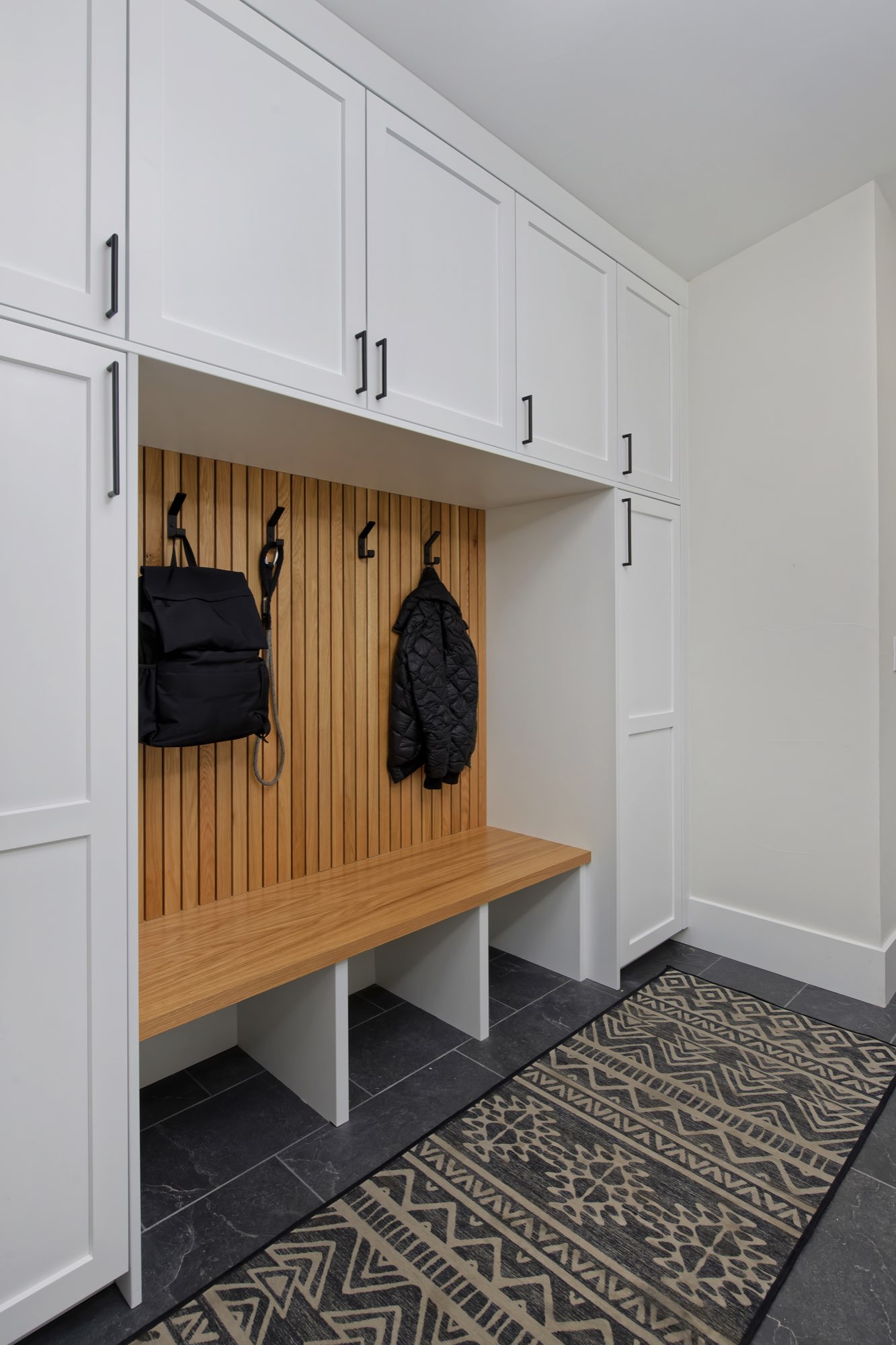 Mudroom with tall white cabinets, a slatted wood panel with hooks, a built-in bench with cubbies and dark tile floor