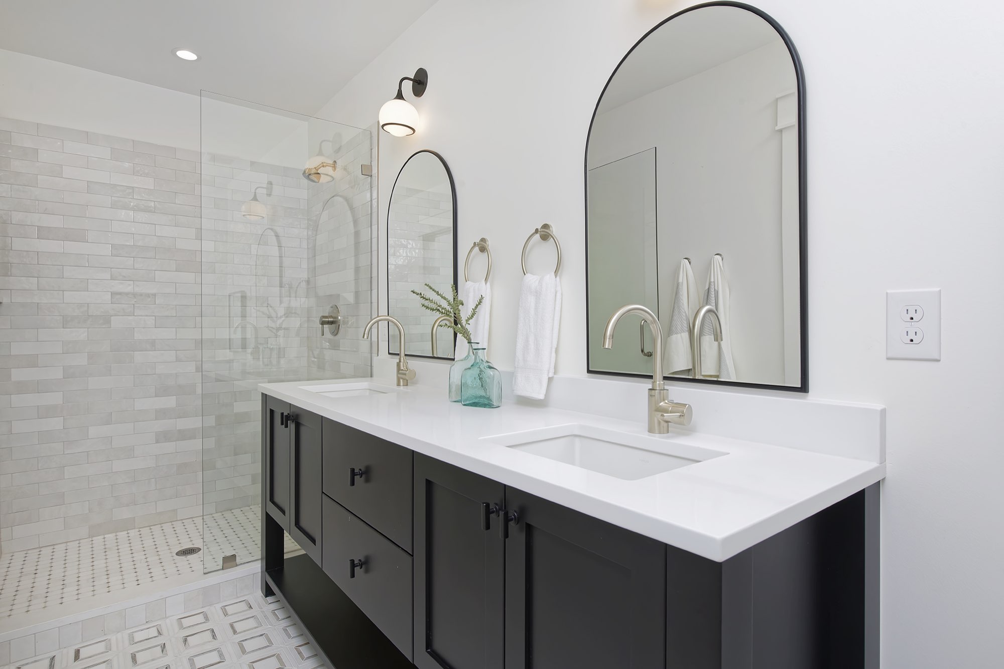 Double black vanity with arched black mirrors, a glass shower and a patterned tile floor