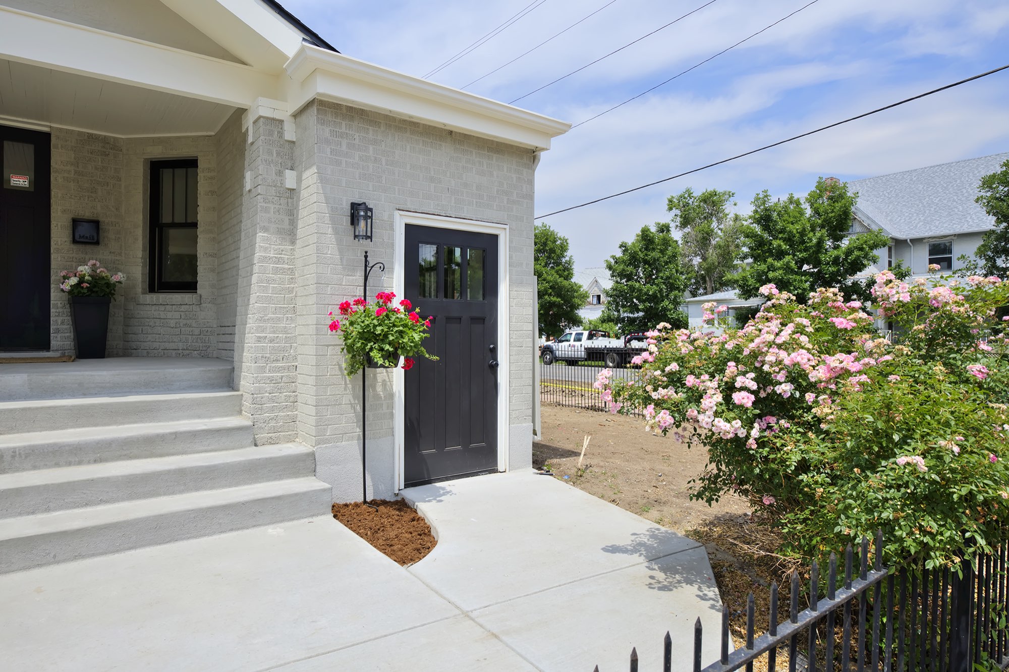 Side entry with a black door, hanging flower basket and concrete steps beside the pale gray brick porch