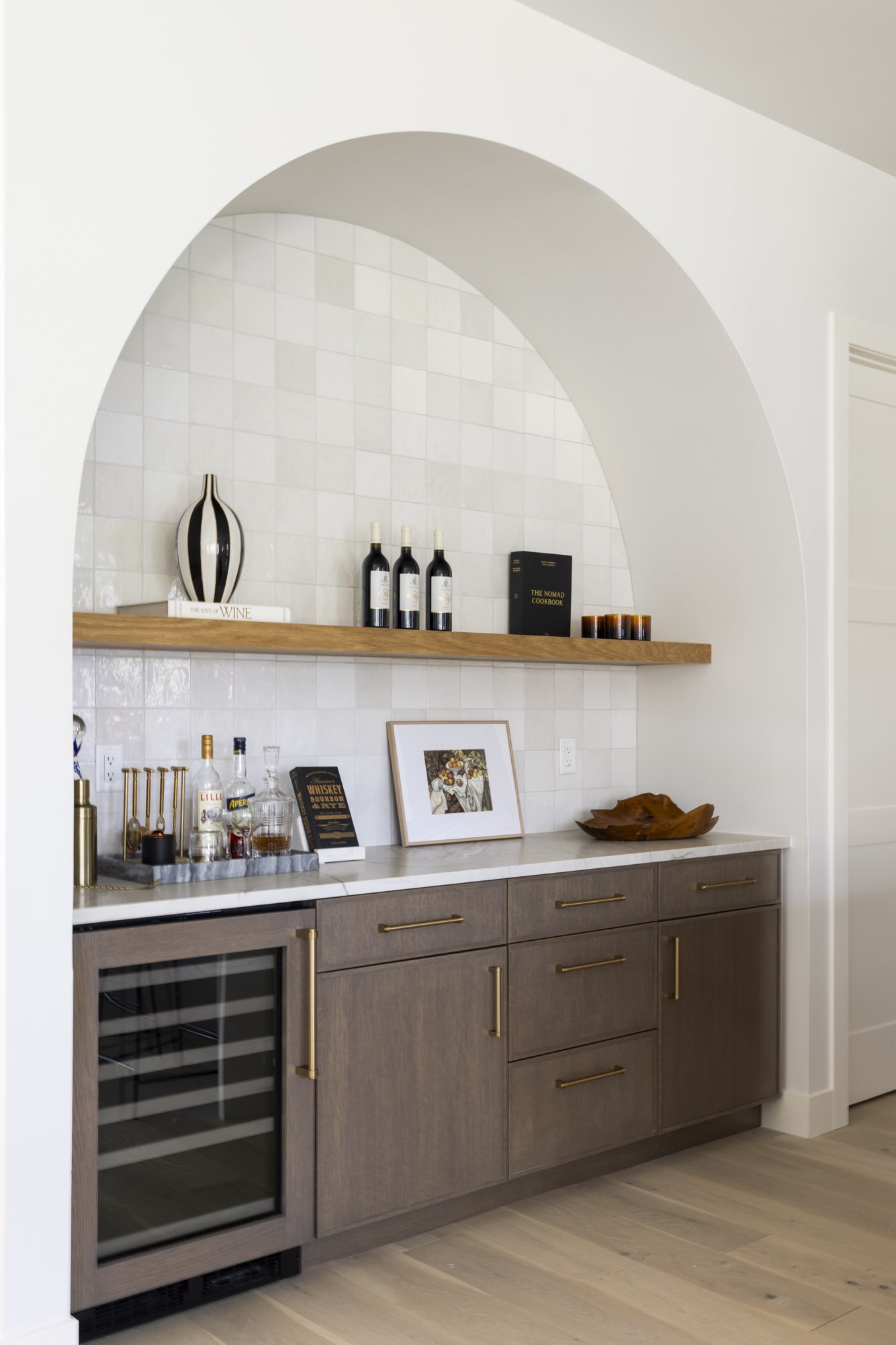 Arched bar niche with a floating wood shelf, light tile, wood-tone cabinets with brass pulls and a wine fridge