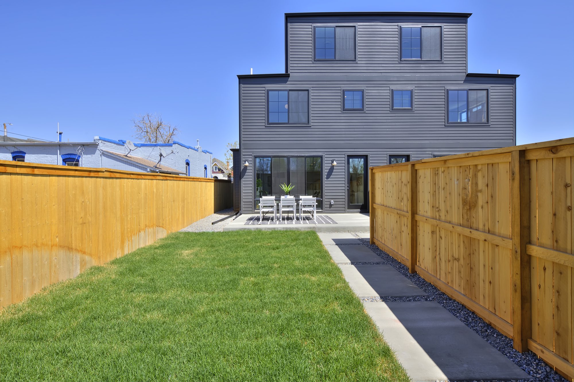 Back of the three-story home in dark siding seen across the lawn, with a concrete walk and a wood fence