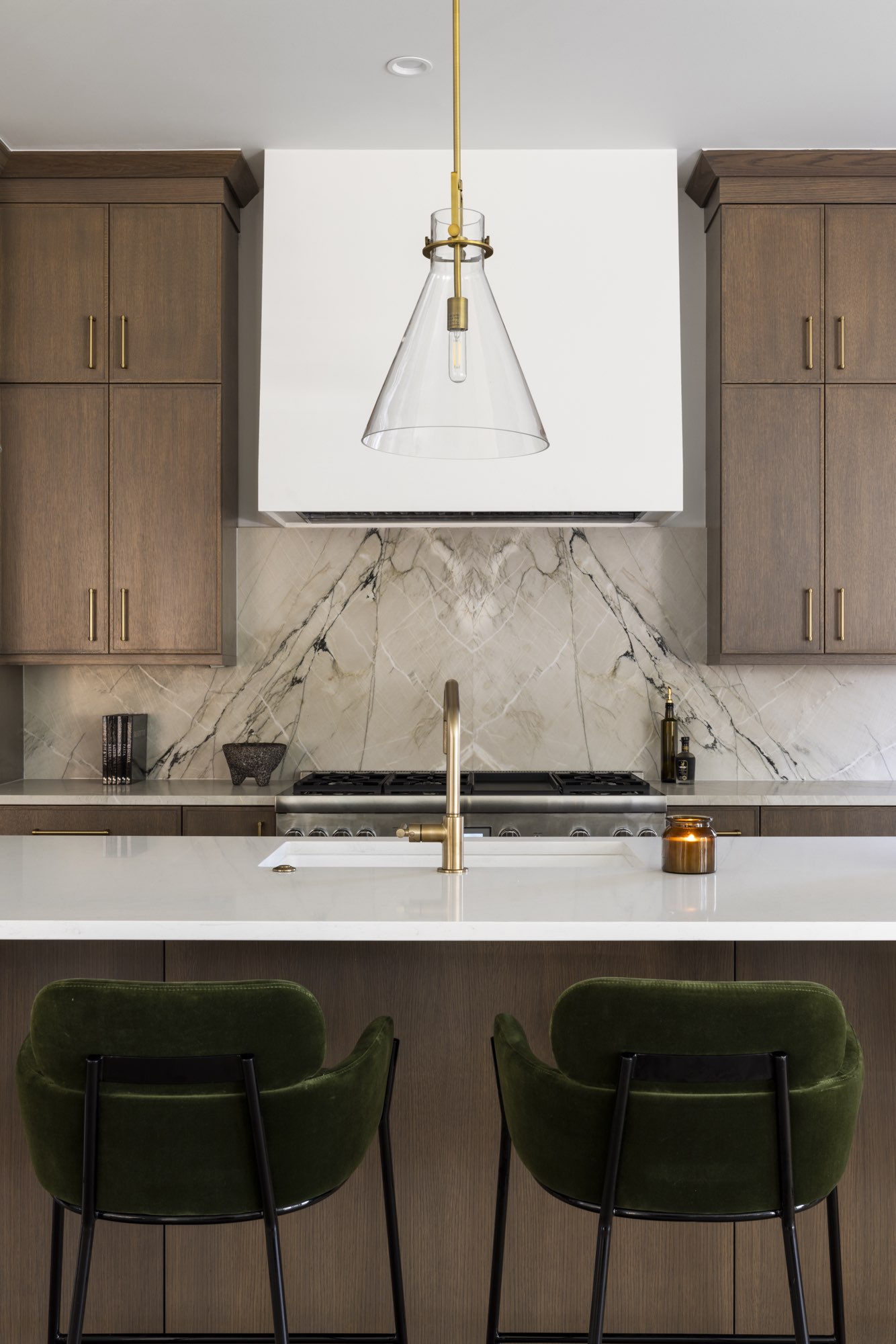 White hood over a range with a veined stone-look backsplash, a brass faucet and a glass pendant