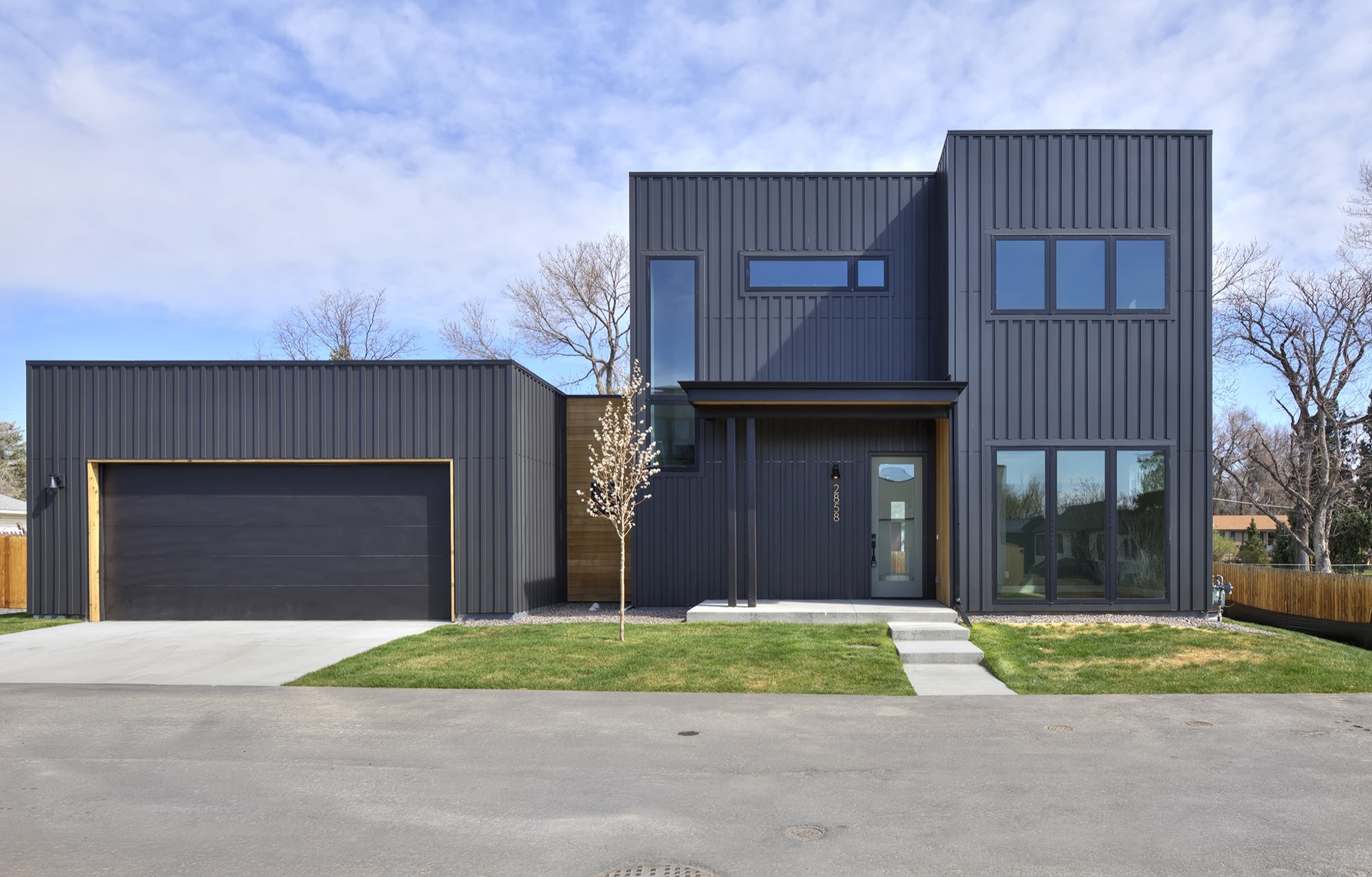 Front of the modern farmhouse at 2858 Teller in Wheat Ridge, with charcoal vertical siding, a wood-trimmed garage door, a pale front door and a young tree in the lawn
