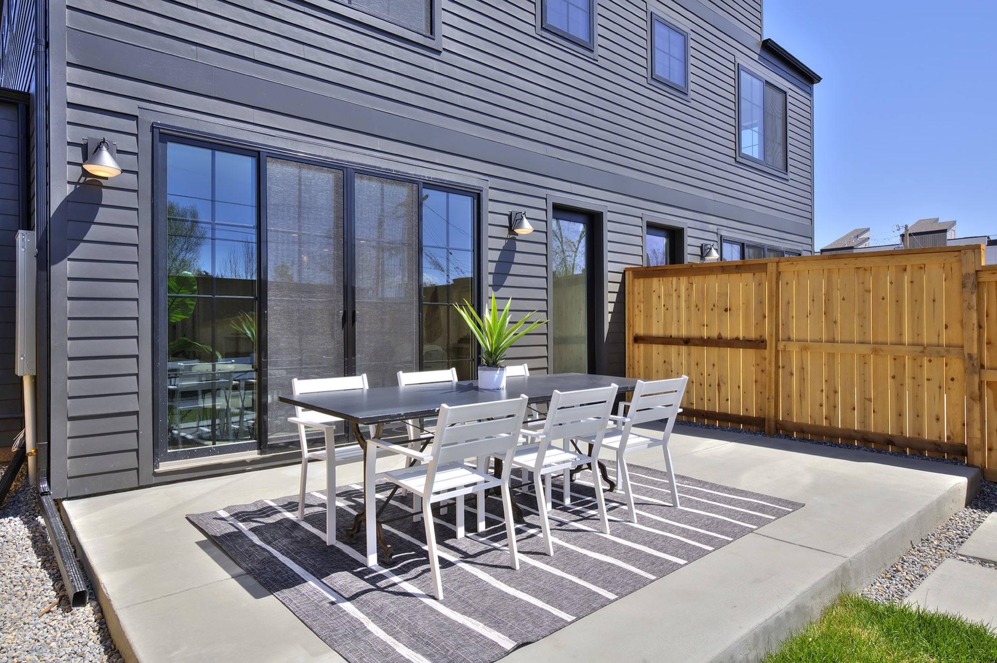 Back of the home in dark siding with black-framed sliding doors, a concrete patio, a dining set and a wood fence
