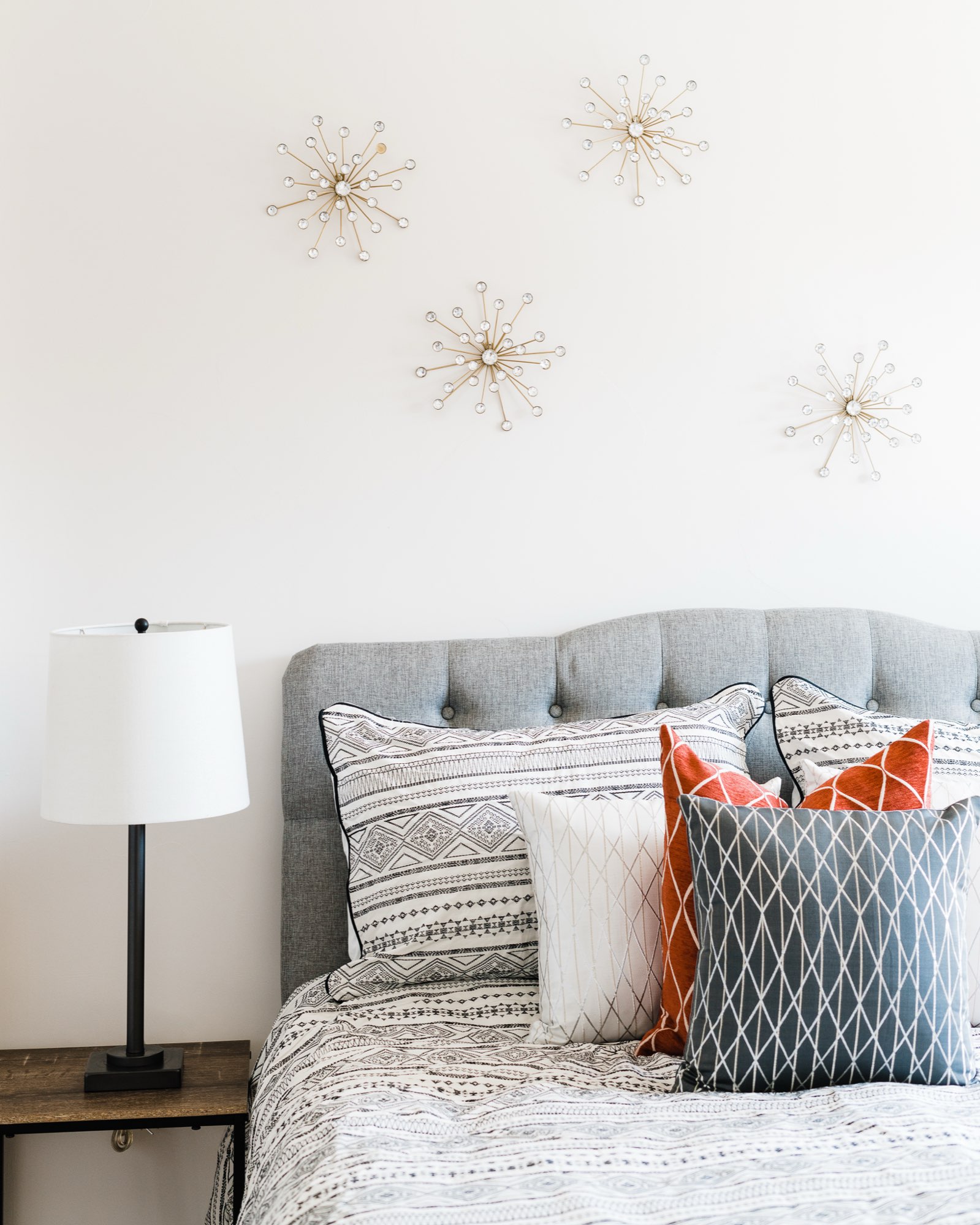 Tufted gray headboard with patterned pillows, a lamp and metal starburst decor on the wall