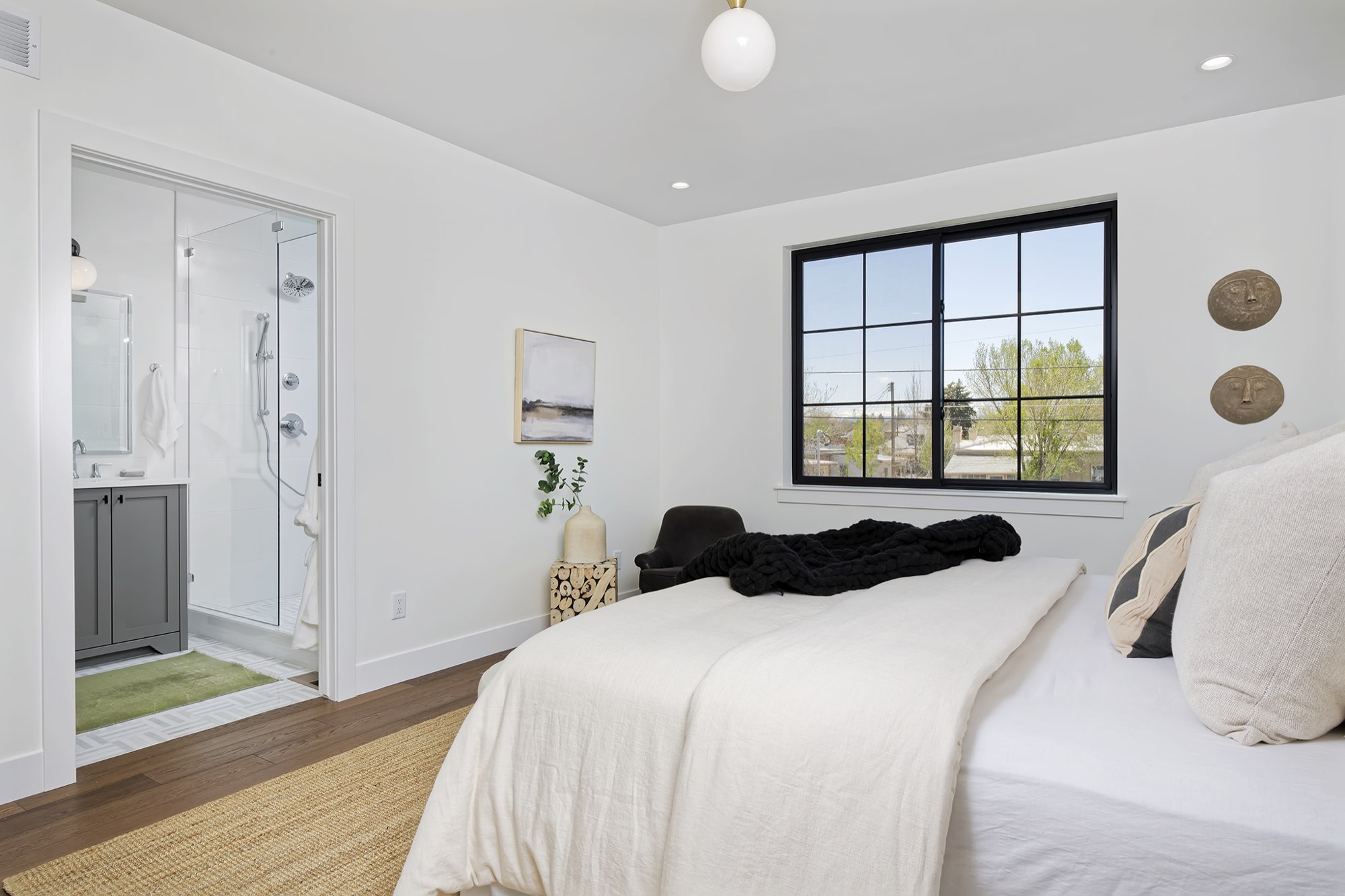 Primary bedroom with a bed, black-framed window, wood floors and a doorway to a bath with a glass shower