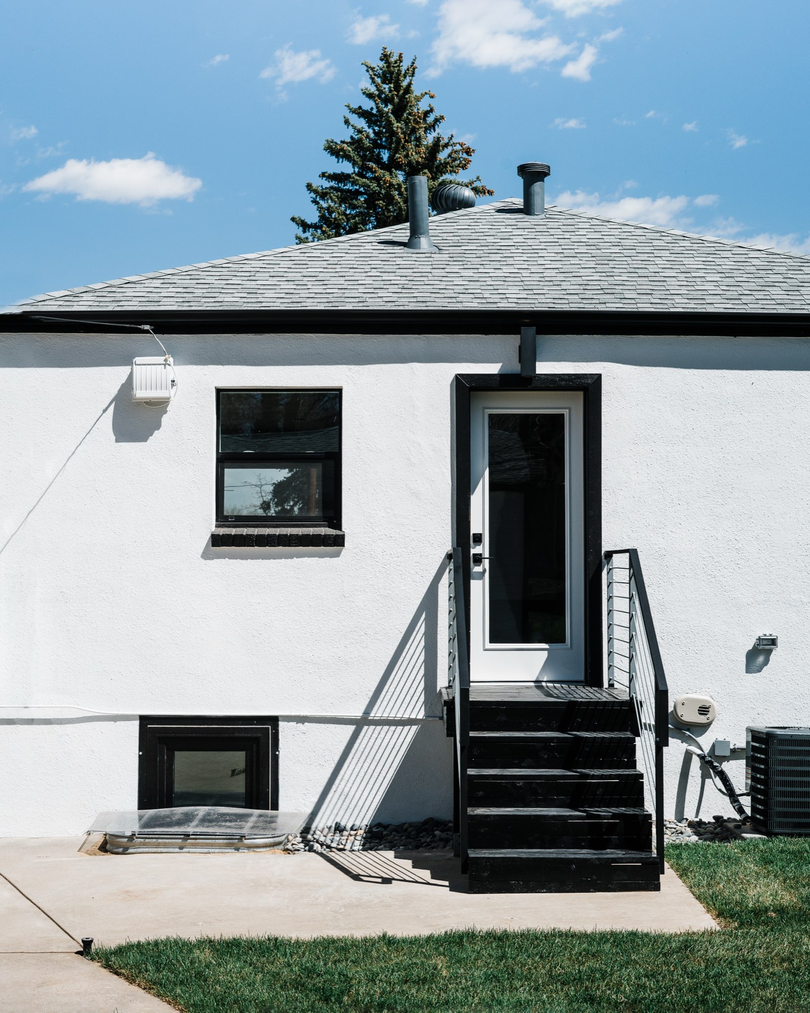 Back of the white bungalow with a white door, black-framed windows, a black stair with railing and a lawn