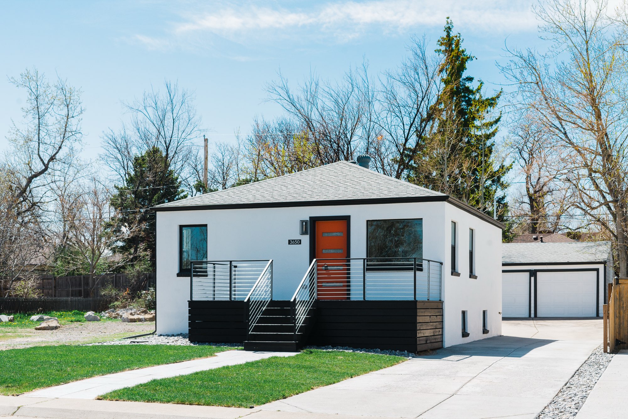Angled front view of the white bungalow with a hip roof, orange door, dark deck and a detached garage at the back of the lot