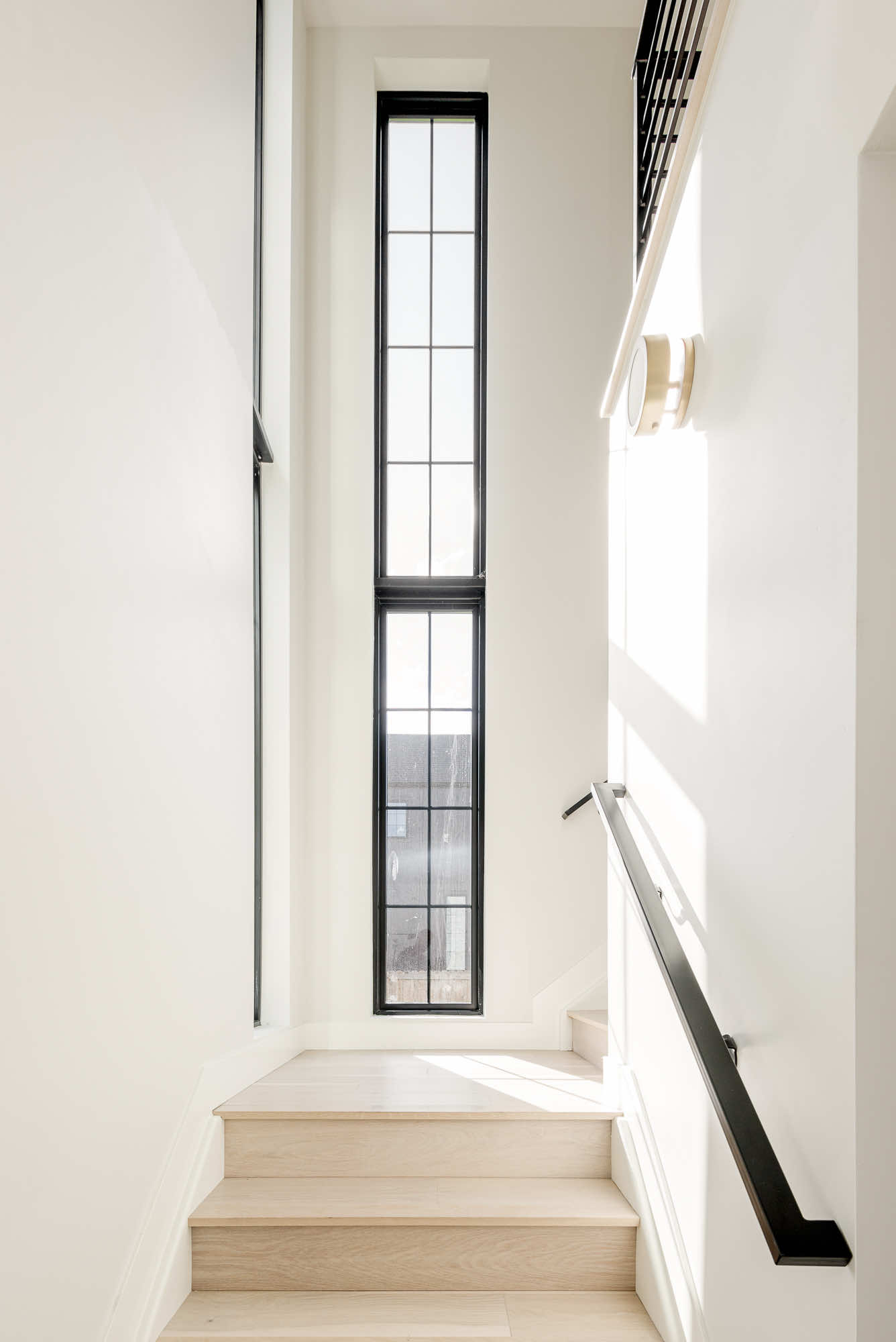 Stairwell with light wood treads, a black handrail and a tall narrow black-framed window